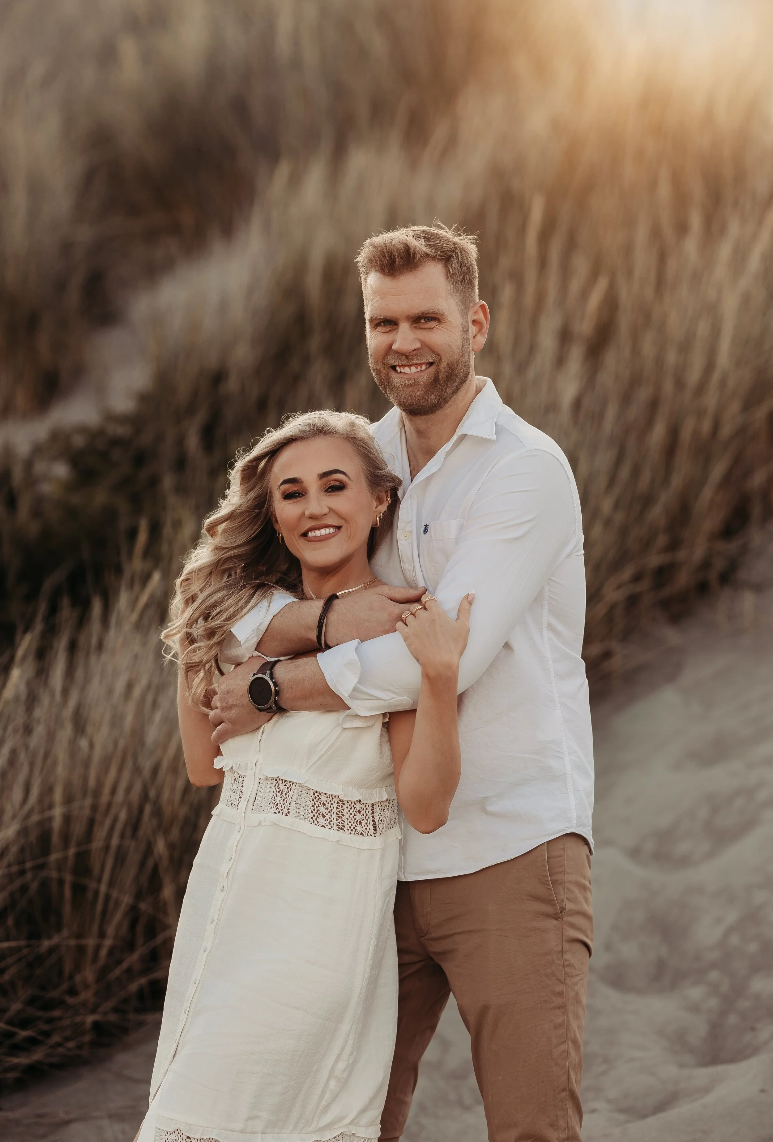 Et smilende par som står nær hverandre på stranden med høye gress i bakgrunnen, kledd i hvite klær.
