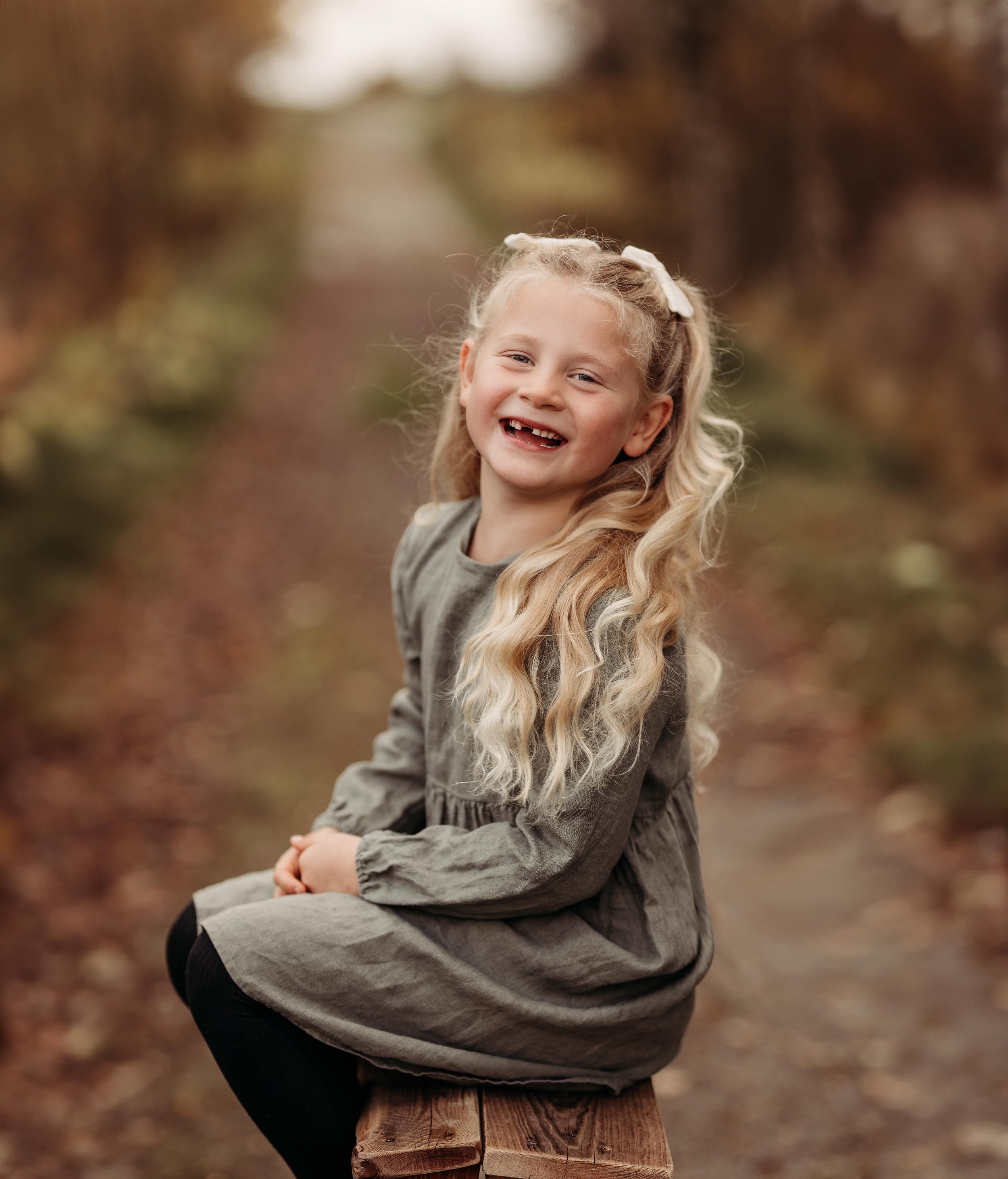 Ei jente med langt, blond, krøllete hår, smilende, sitter på en trestamme i en skog, høstfarger i bakgrunnen.