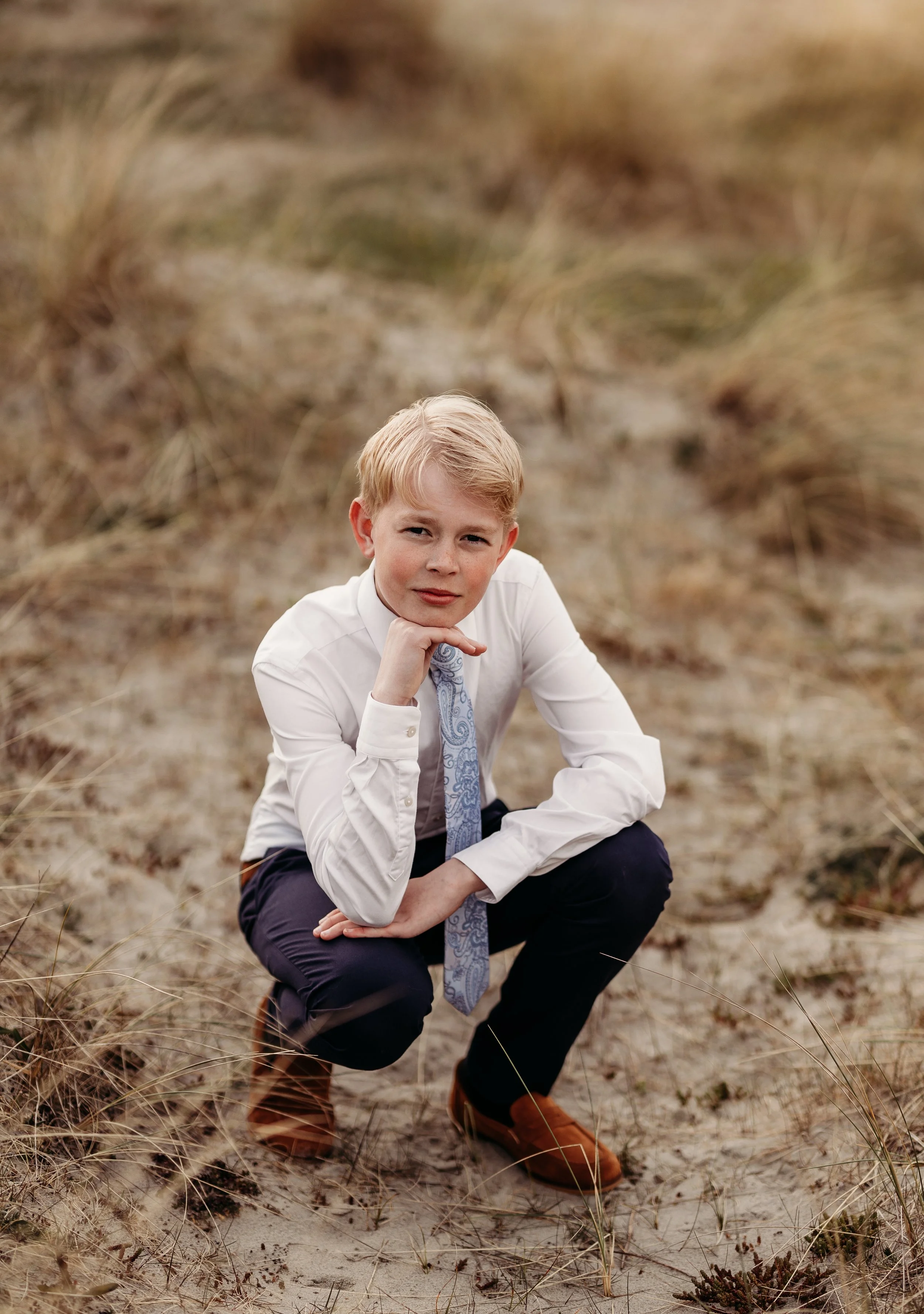 Ung mann i dress og slips, legger en hånd under haken, knelende på sanddyne med gress, ser direkte i kamera.