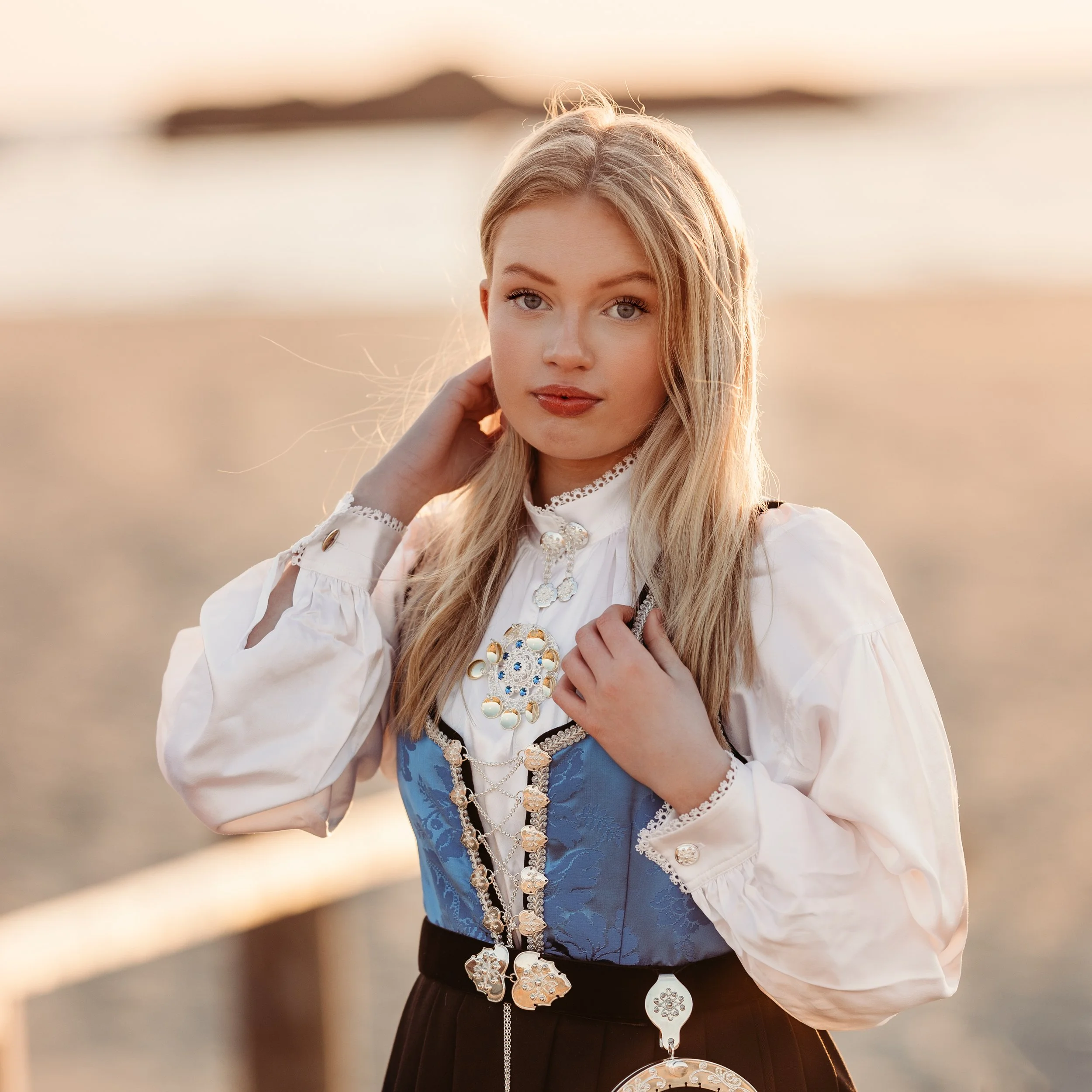 En ung kvinne i tradisjonell norsk bunad står ved en strand under solnedgang.