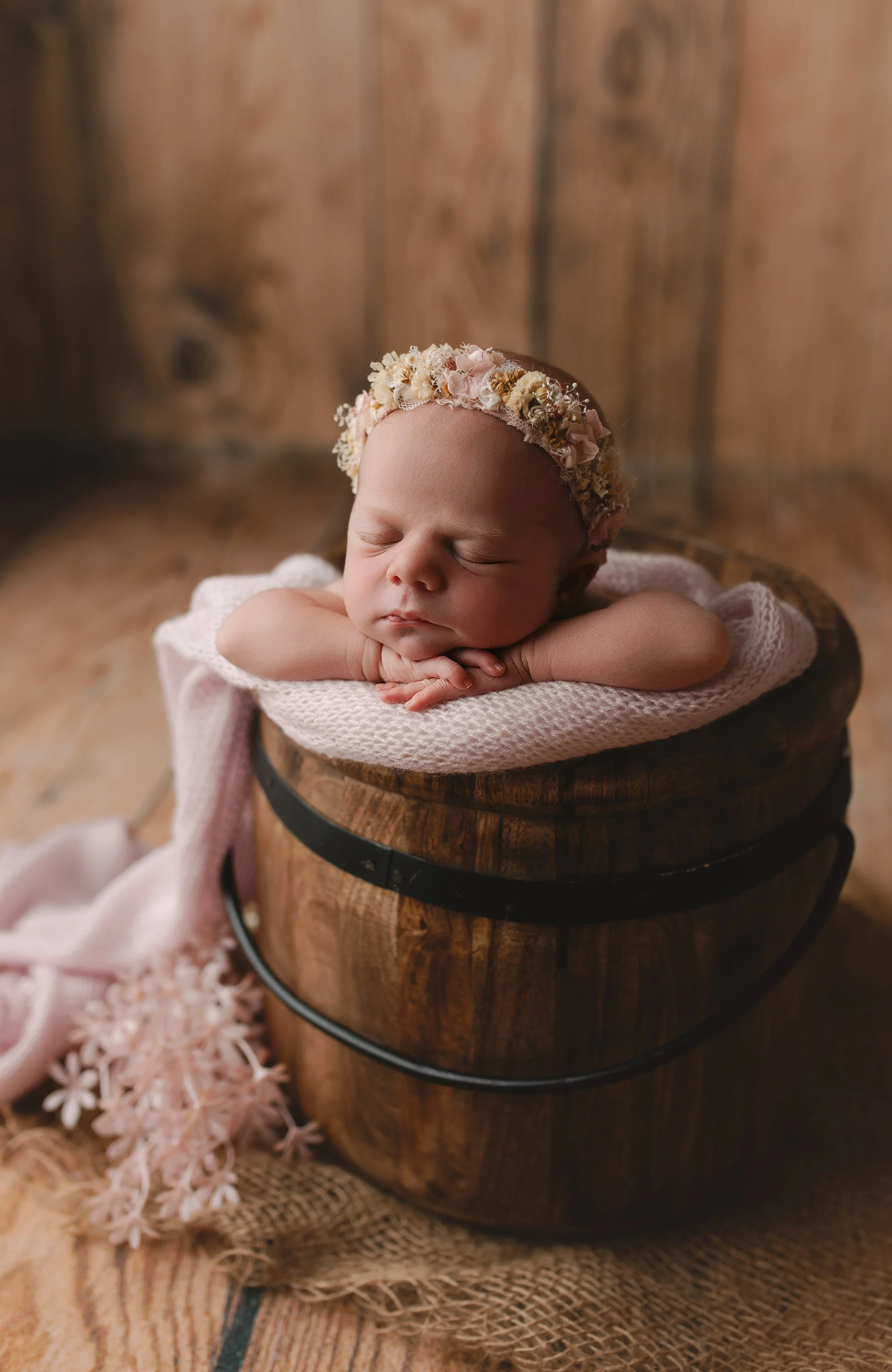 Søvnende baby med blomsterhodebånd, liggende på armene oppå en trekasse, omgitt av treverk og mykt stoff.
