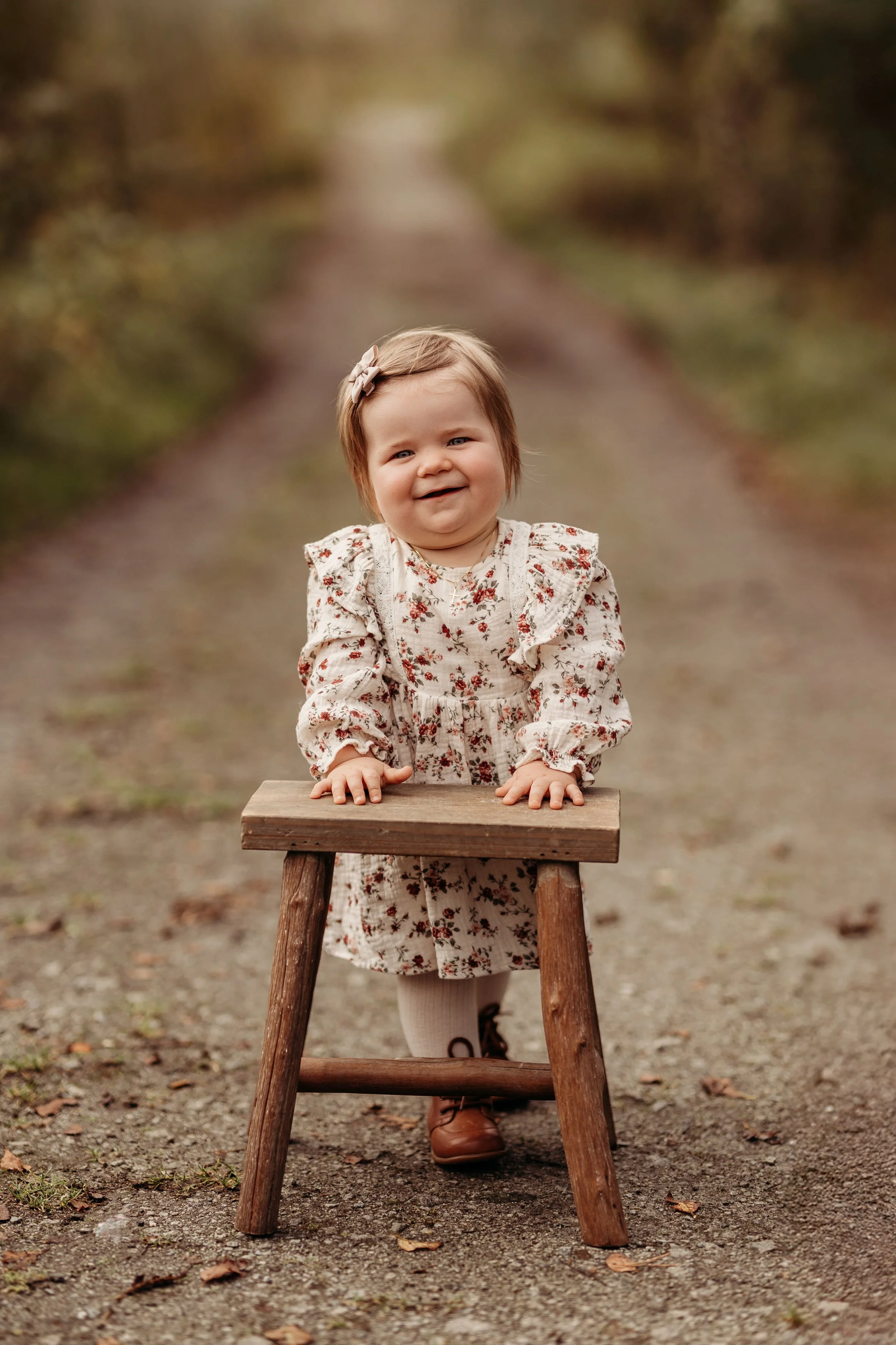 En liten jente med kort hår og en blomsterkjole står bak en treskrakk på en sti i en skog. Hun smiler og bøyer seg litt fremover, med hendene på skranken.