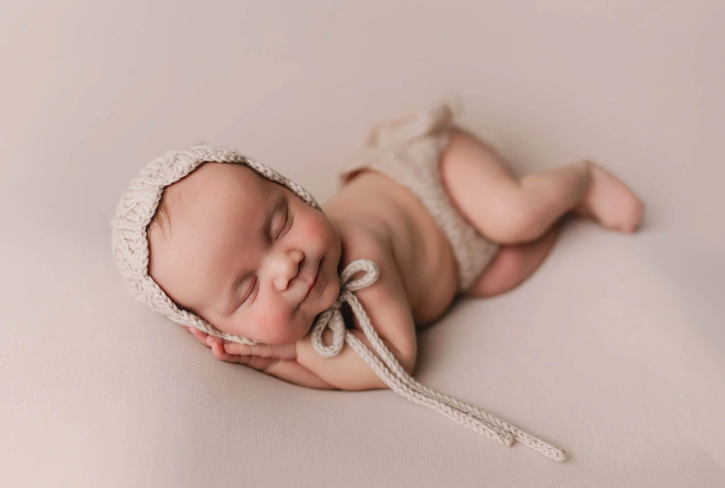 Søvnende nyfødt baby med strikket lue og bleie, liggende på mage på nøytral farget bakgrunn.