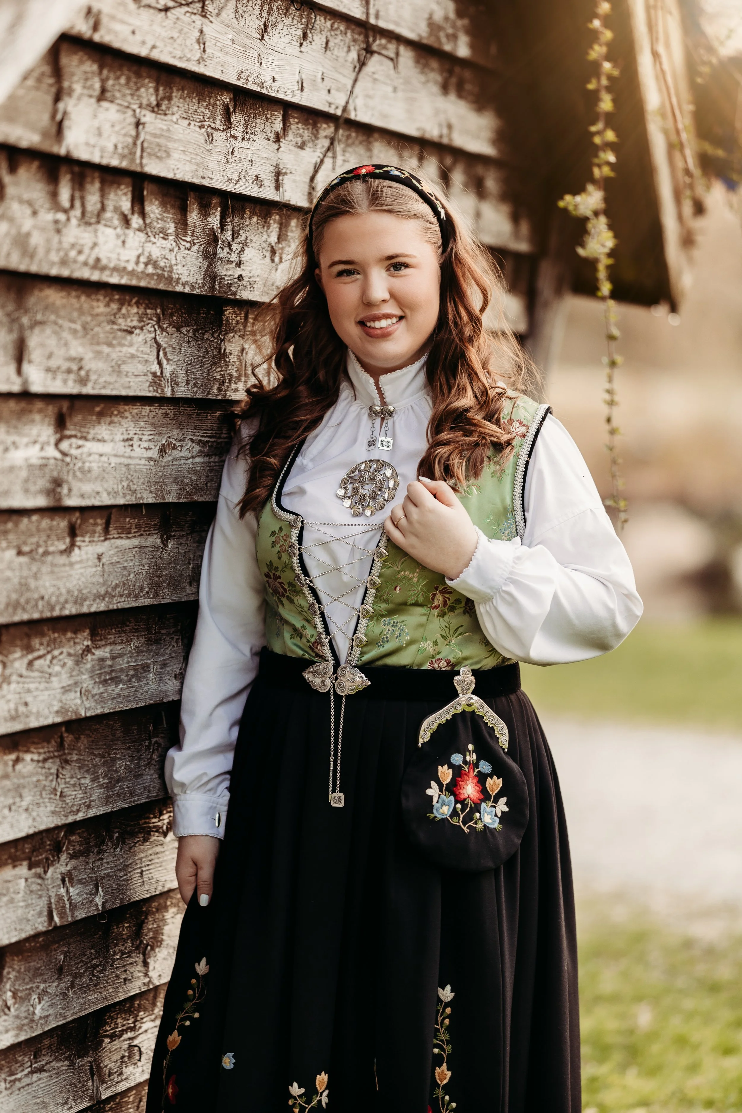 En jente kledd i tradisjonell norsk bunad står ved en trevegg. Hun smiler og har langt, krøllet brunt hår og en blomsterdekorert hårbøyle.