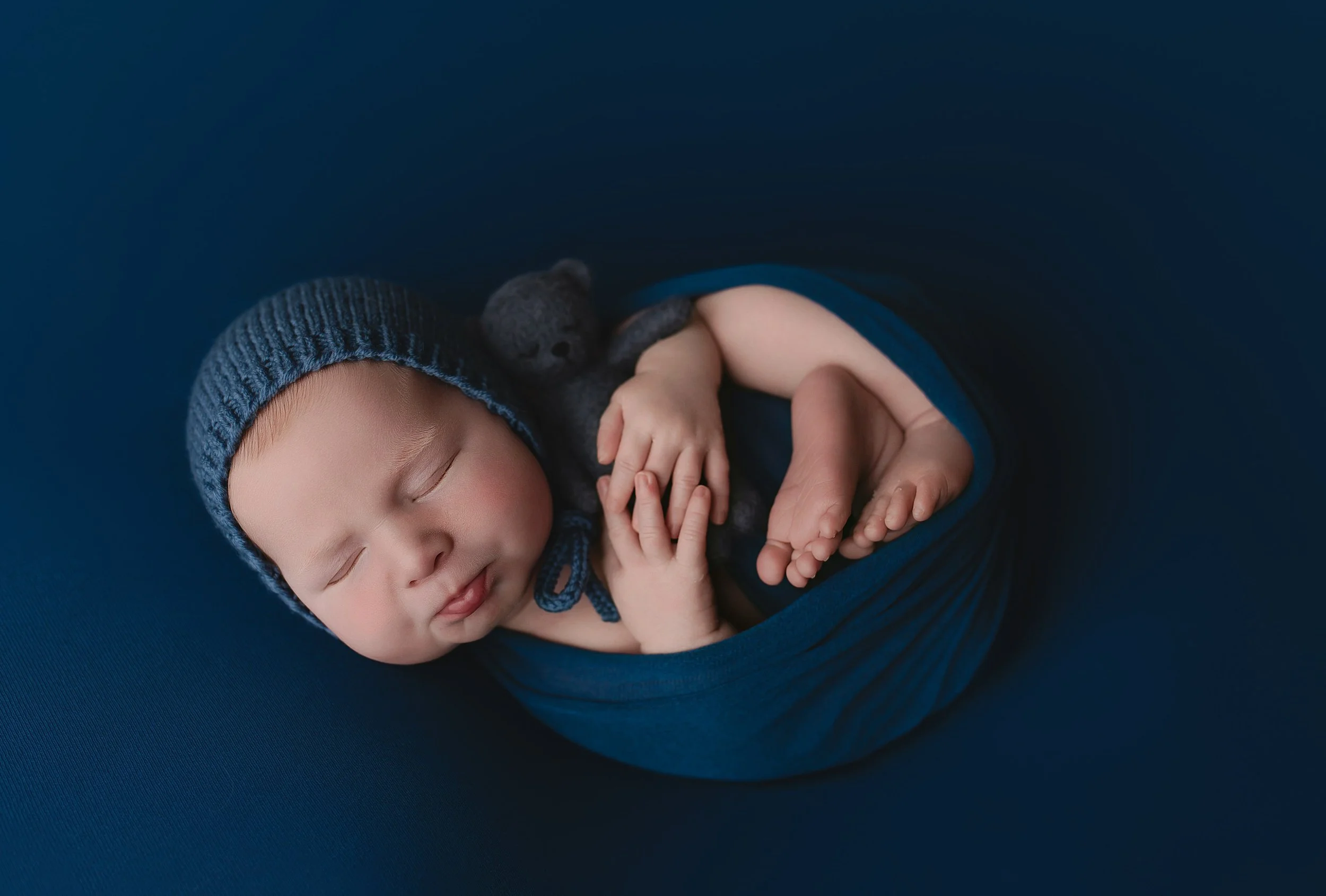 Sovende baby med pysjamas og strikket lue, rørende klem med en liten svart bamse på en mørk blå bakgrunn.