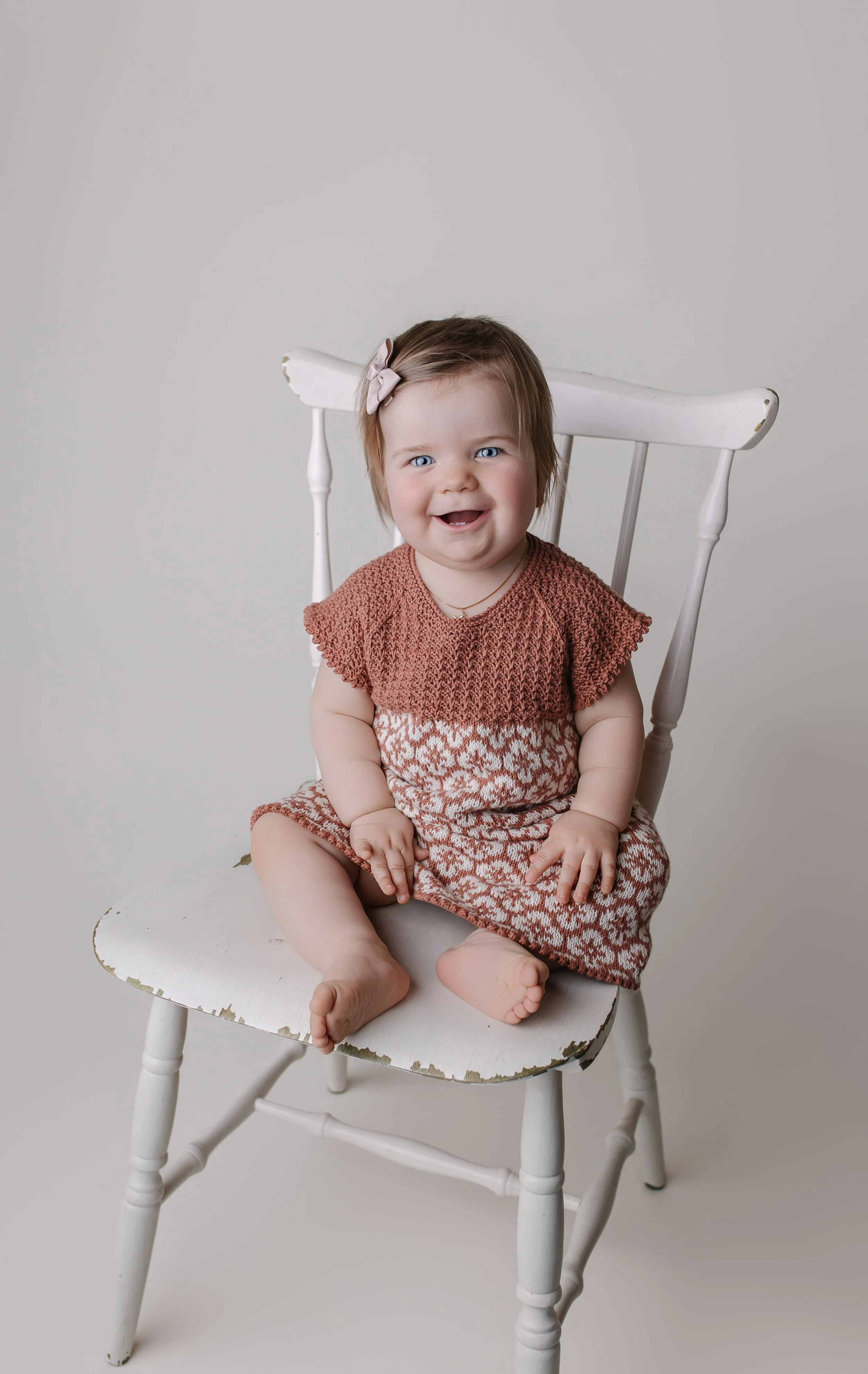 En liten jente med brunt hår og blå øyne, sitter på en gammel hvit stol mot en lys bakgrunn. Hun smiler og ser rett mot kamera, og har på seg en rosa og hvit kjole med en lys rosa hårspenne.