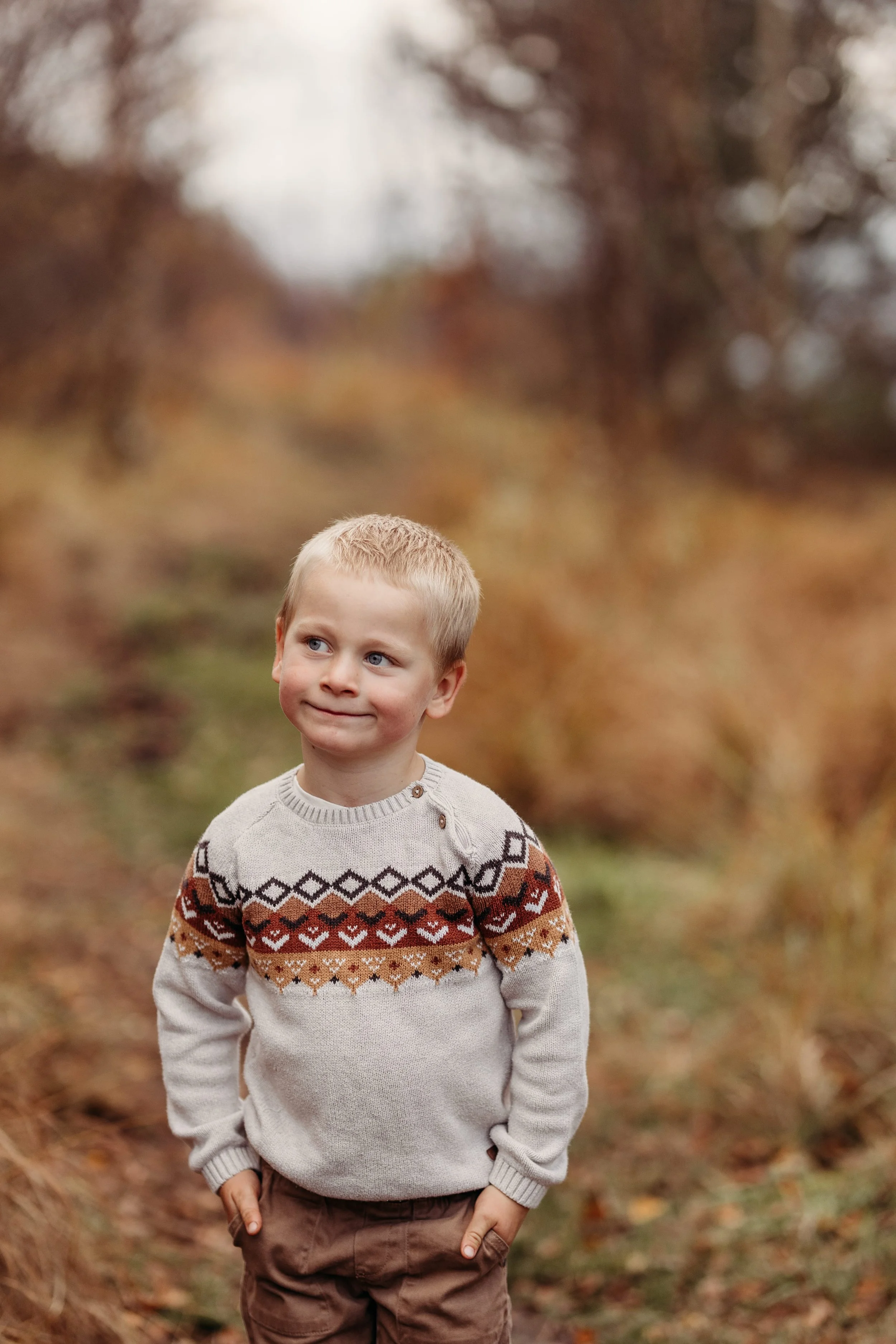 En ung gutt står på en sti i en skog med høstfarger, iført en lys genser med mønster og brune bukser.