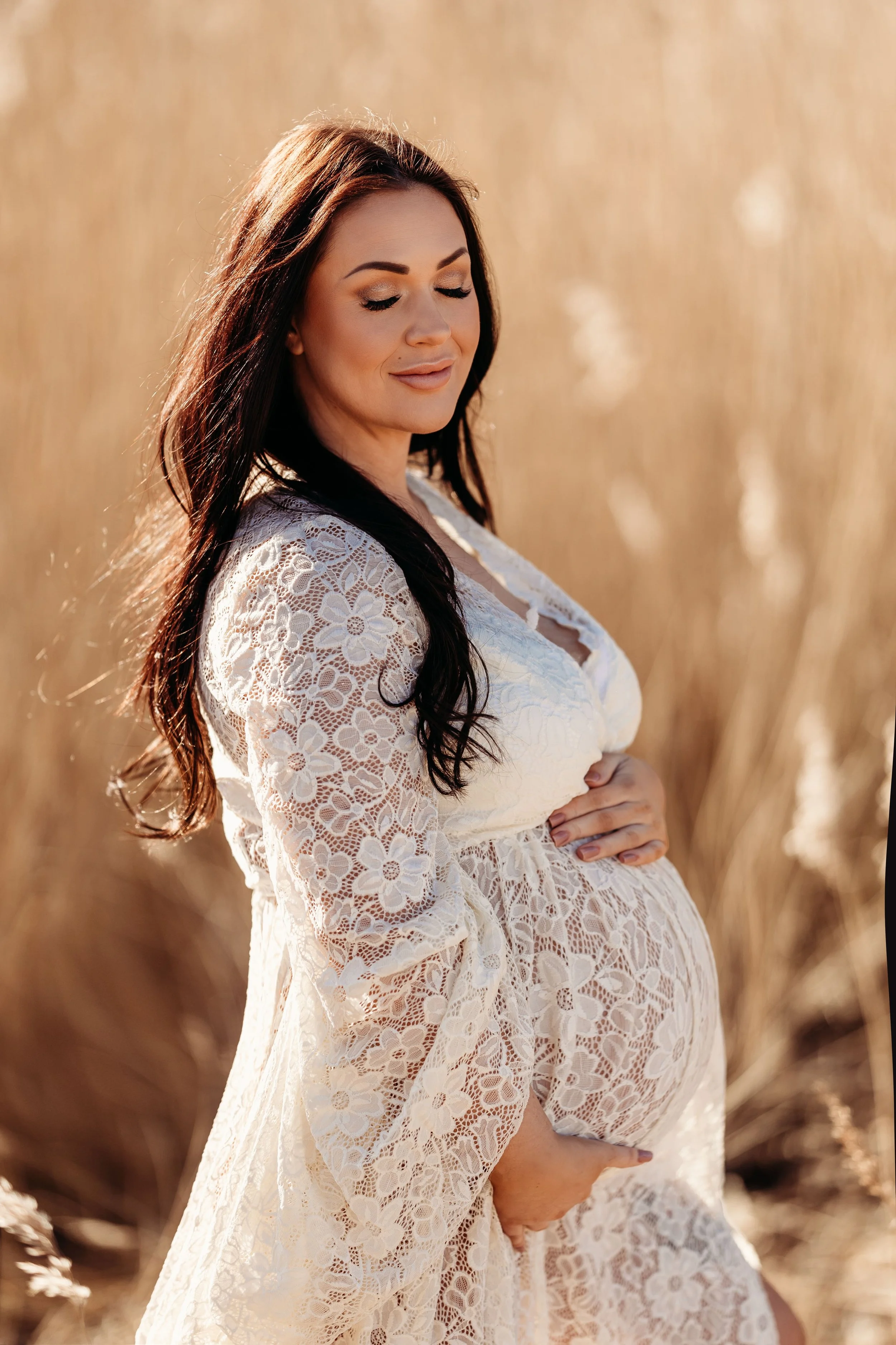 Gravid kvinne i hvit blonderkle ferdig med å rose magen sin i felt med høyt gress, solskinn.
