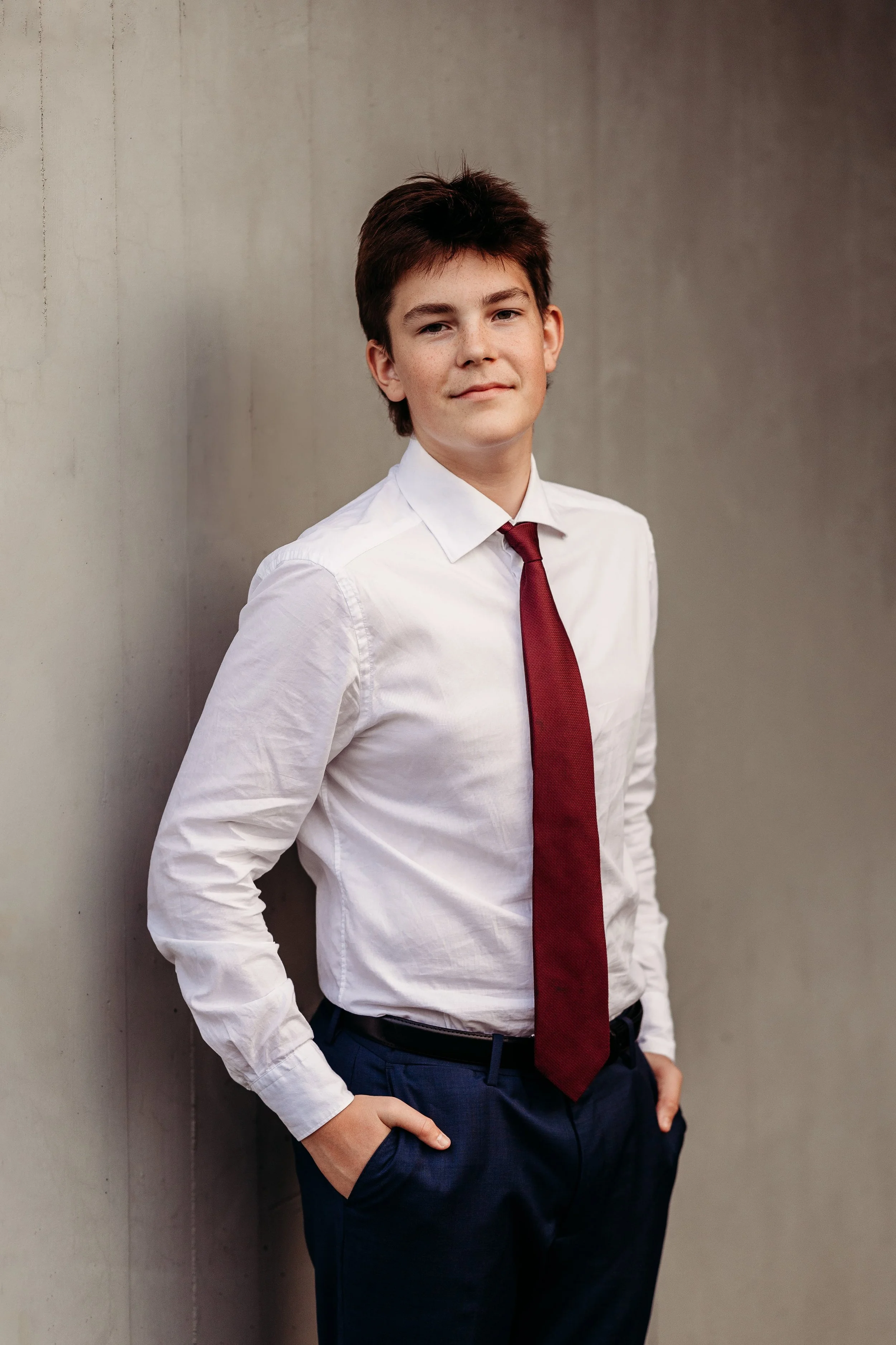 En ung mann iført hvit skjorte, rød slips og blå bukser, står med hendene i lommer ved en grå vegg, ser rett inn i kamera.
