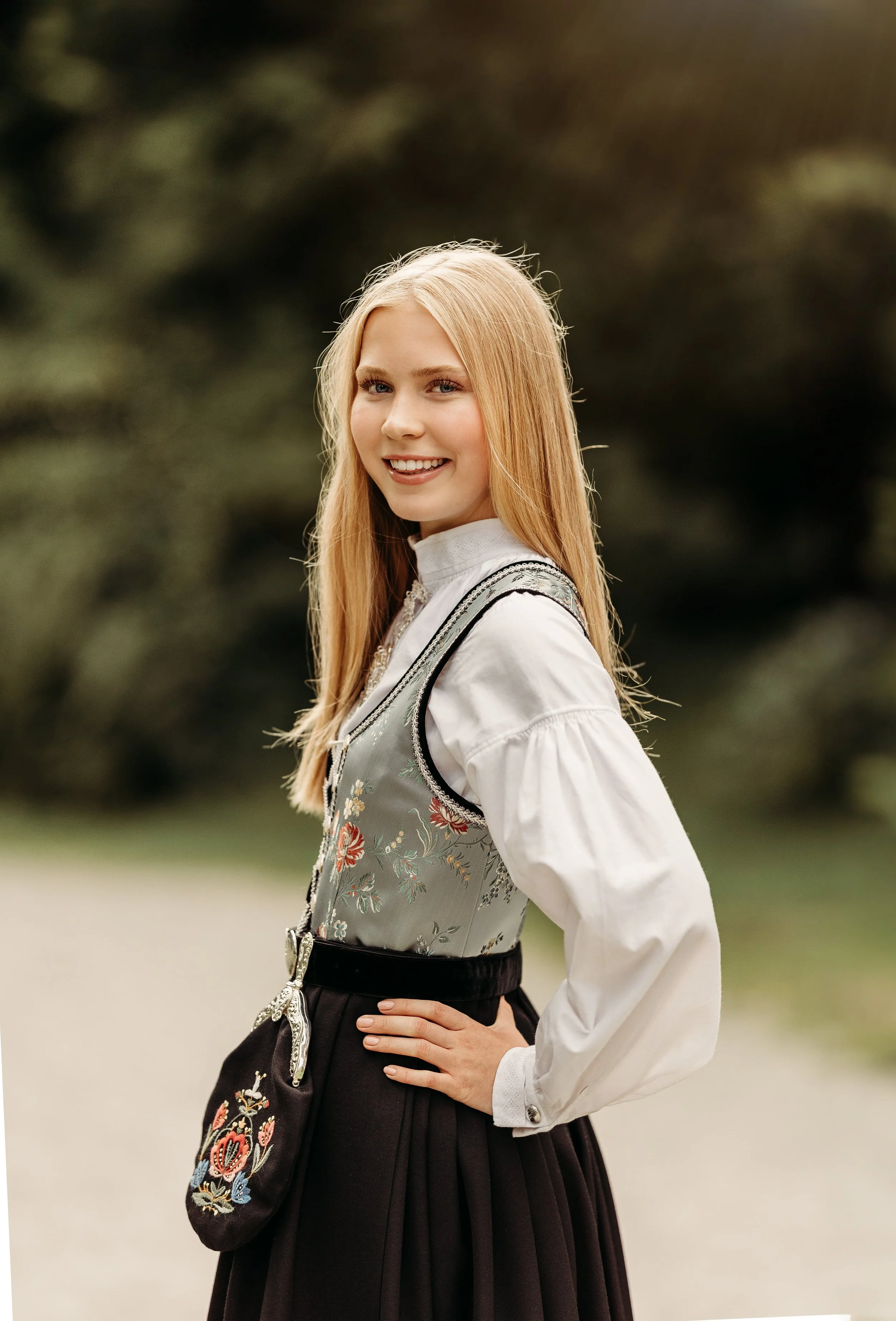 En ung kvinne smiler og ser mot kamera, iført tradisjonell norsk bunad med intrikate broderier, i utendørs omgivelser med grønne trær i bakgrunnen.