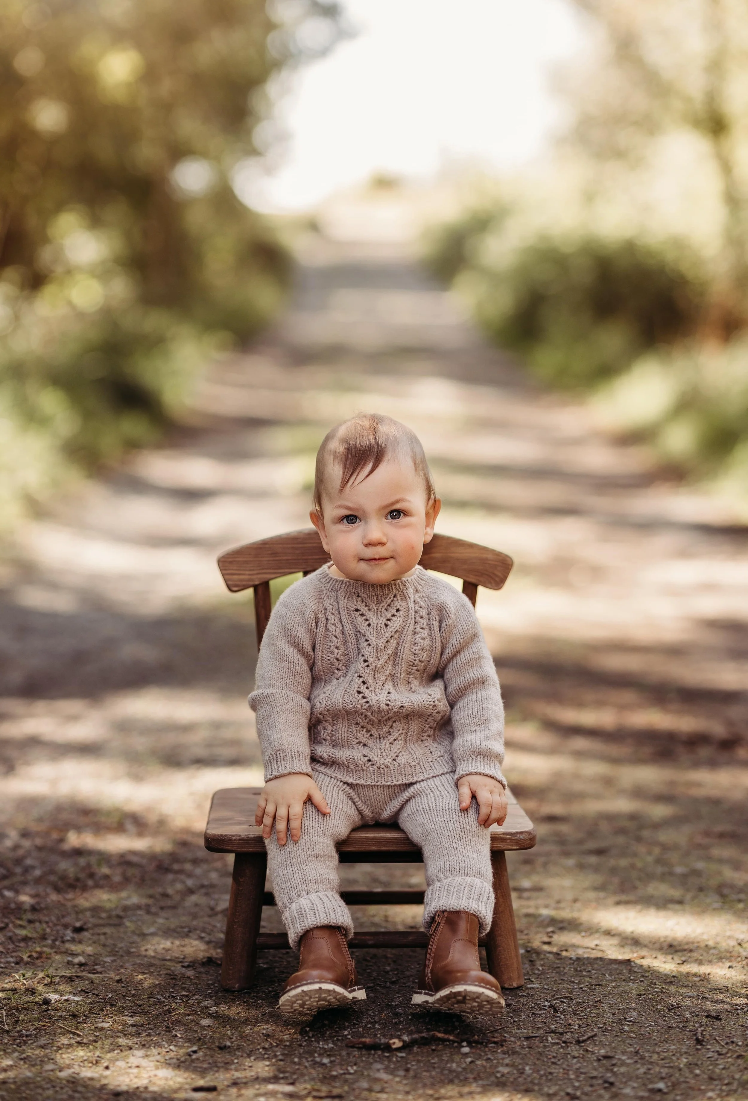 En liten gutt i beige klær og støvler sitter på en trebue i en skogsti.