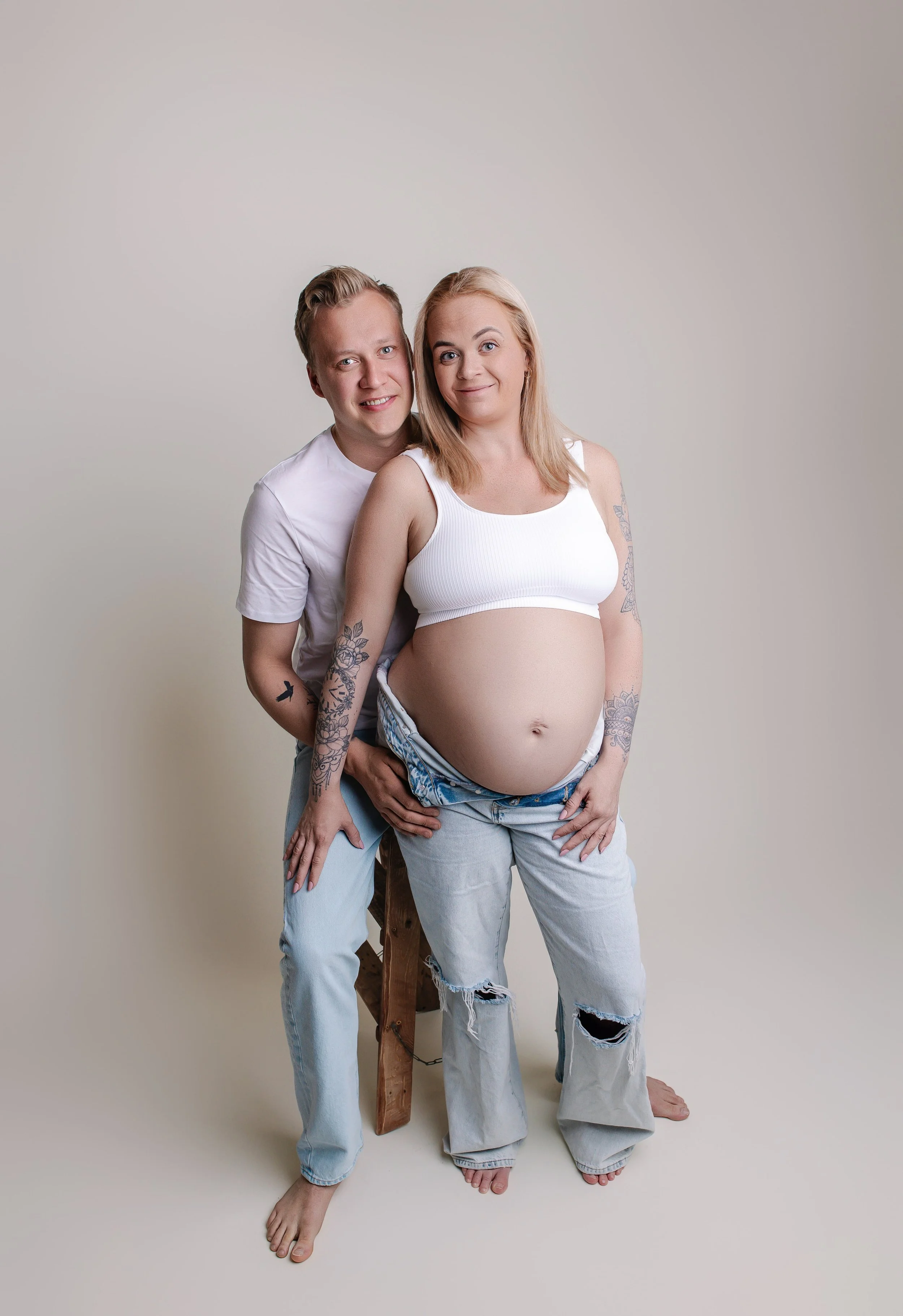 Et ungt par og en gravid kvinne poserer sammen mot en lys, enkel bakgrunn. Kvinnen har langt blondt hår, tatoveringer på armene, og har på seg topp og løse jeans. Mannen står bak henne, holder rundt hoften hennes og smiler.