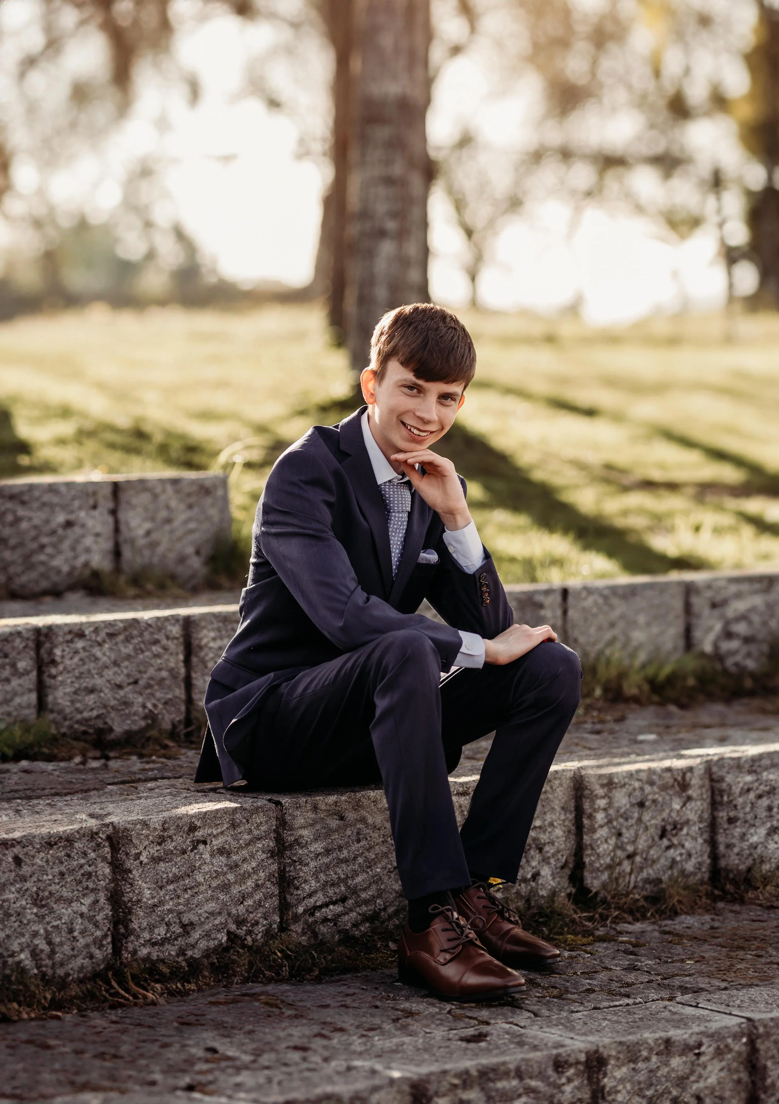 En ung mann i dress sitter på trappen utendørs, sol og trær i bakgrunnen.