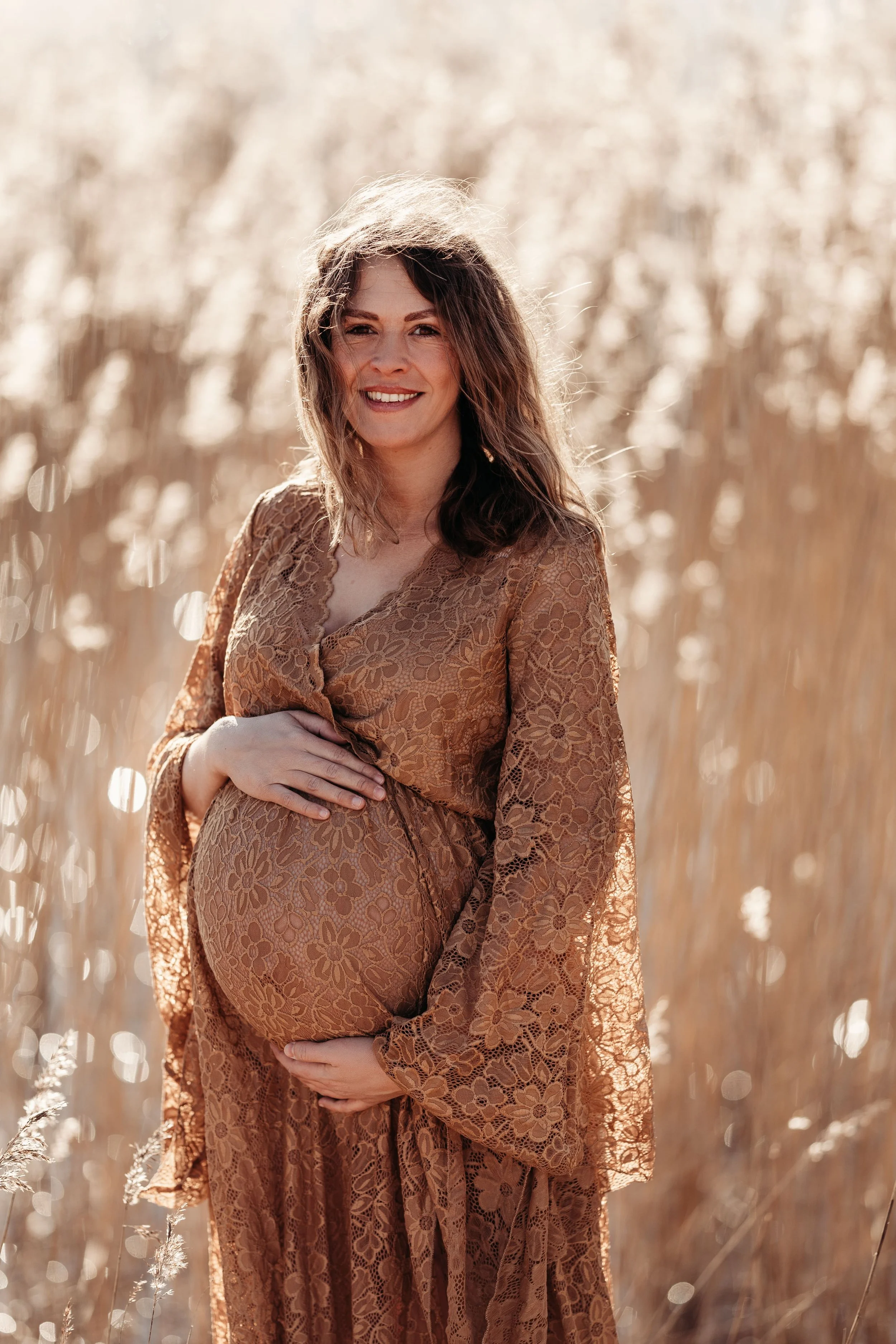 Gravid kvinne i brunt blonder kjole står i en åker med høstfarger, smiler og holder magen med begge hender.