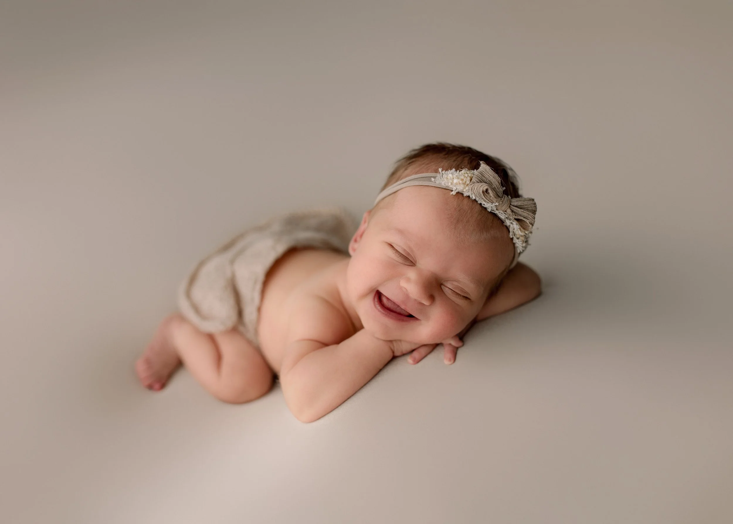 En baby ligger på magen, smilende og med lukket øyne, iført en beige klær og en blomstrete hodebånd, på en lys bakgrunn.