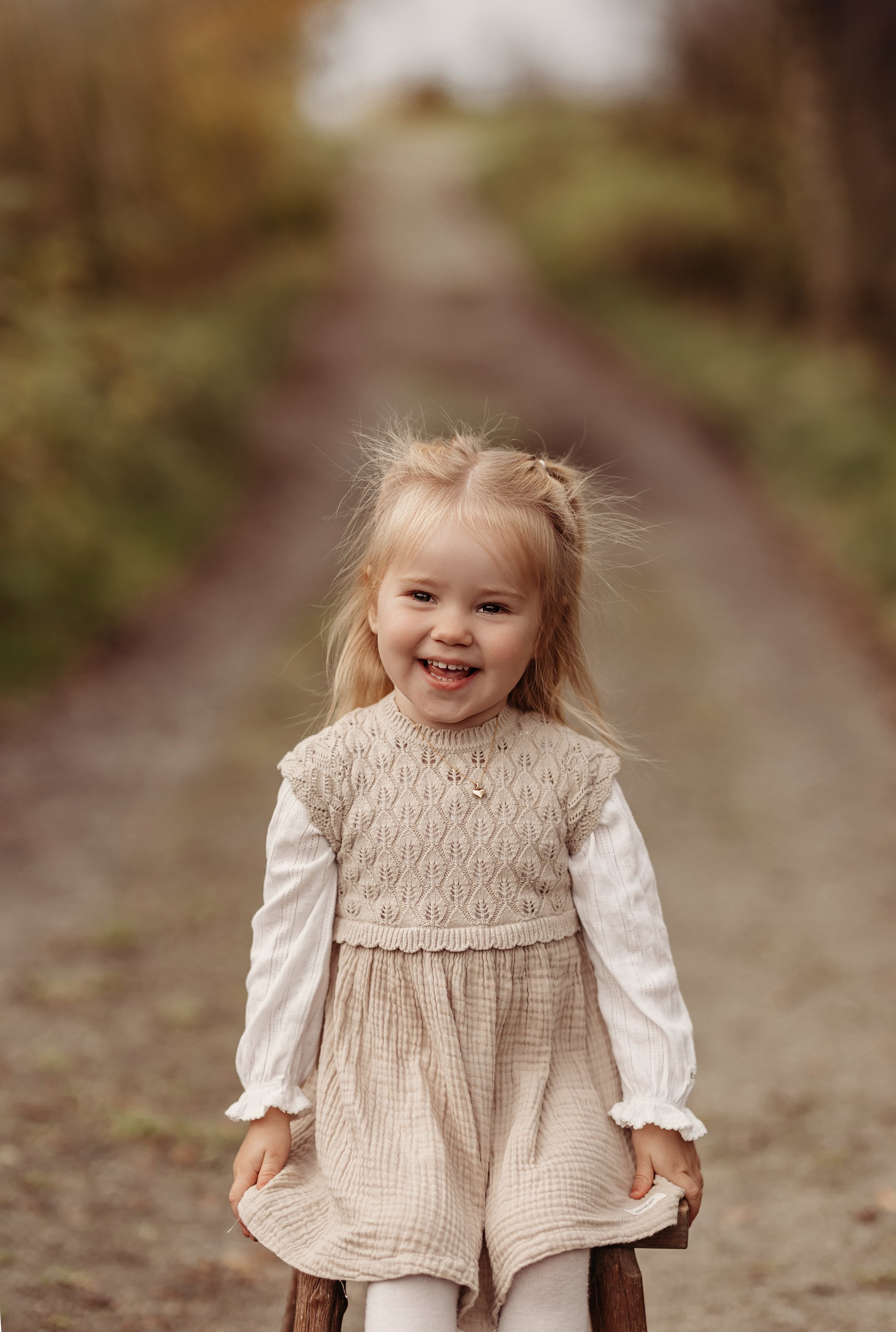 Liten jente med blondt hår som smiler, sitter på en trebrygge på en skogssti.