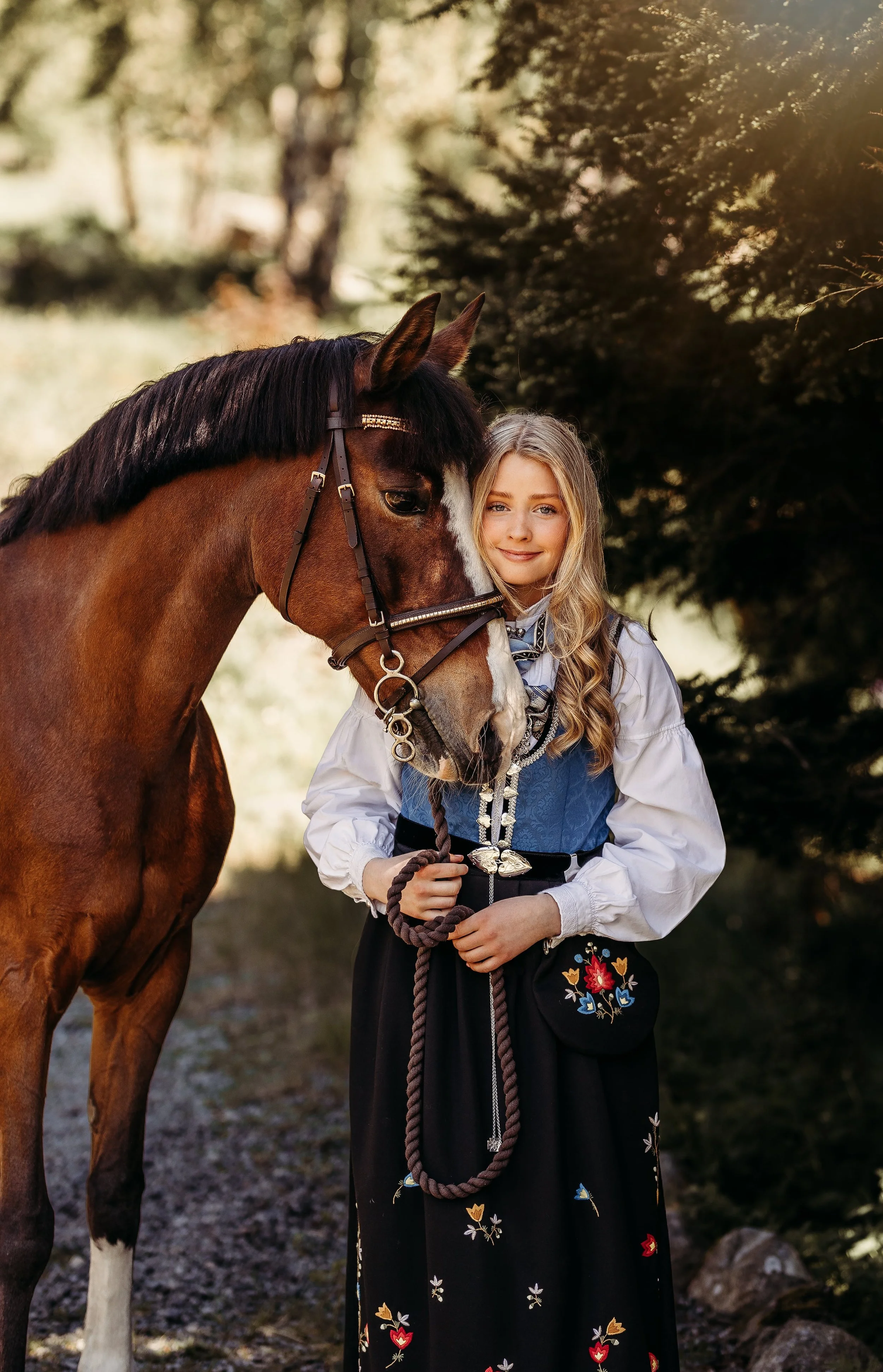 En jente i tradisjonell norsk folkedrakt med en brun hest utendørs på dagtid med grønt løvverk i bakgrunn.
