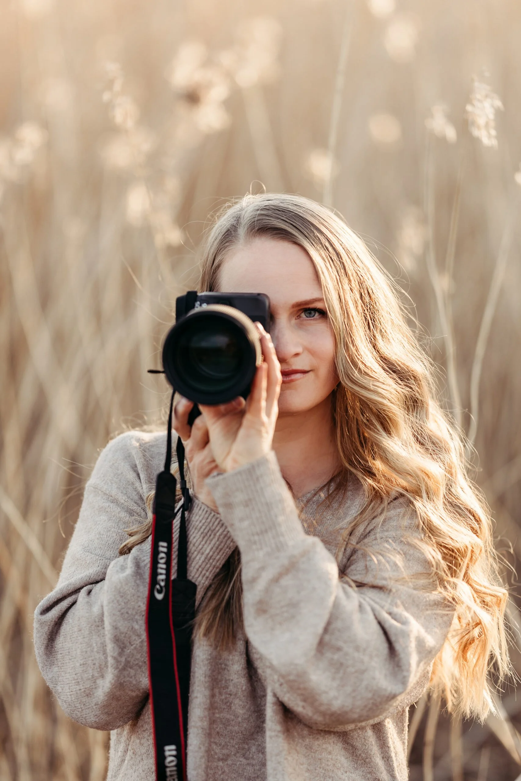 En kvinne med langt, blondt, krøllet hår som tar et bilde med et DSLR-kamera utendørs i en åpen felt med tørt gress.