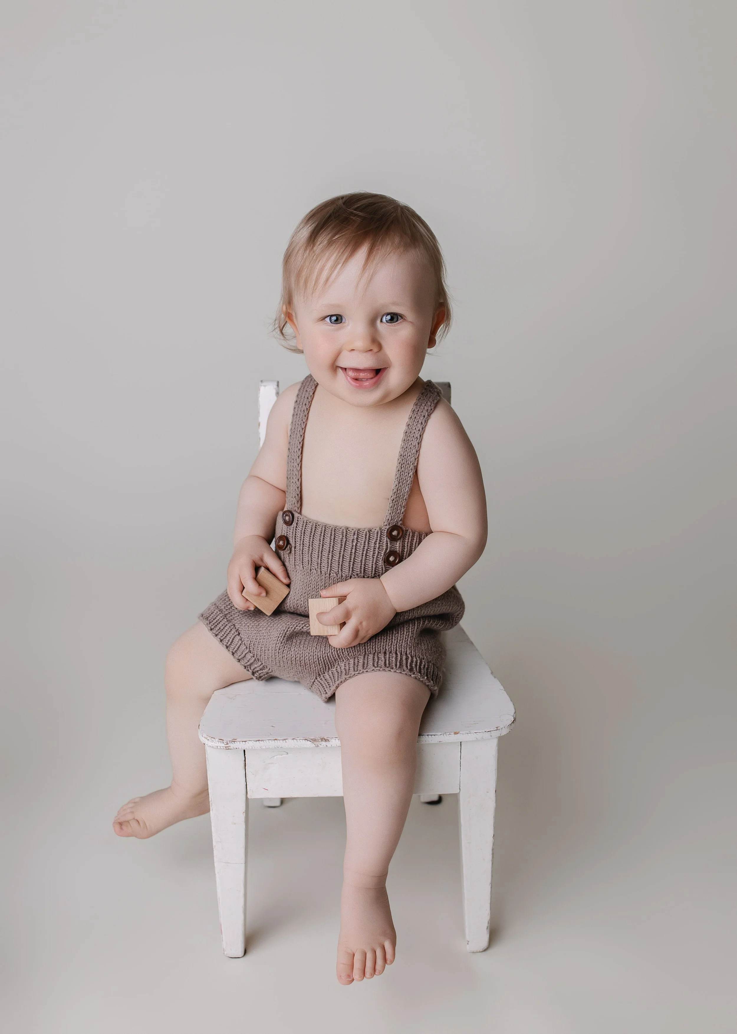 En smilende barn med blond hår, sitte på en hvit trestol, iført beige strikkede bukser og uten skjorte, mot en lys bakgrunn.
