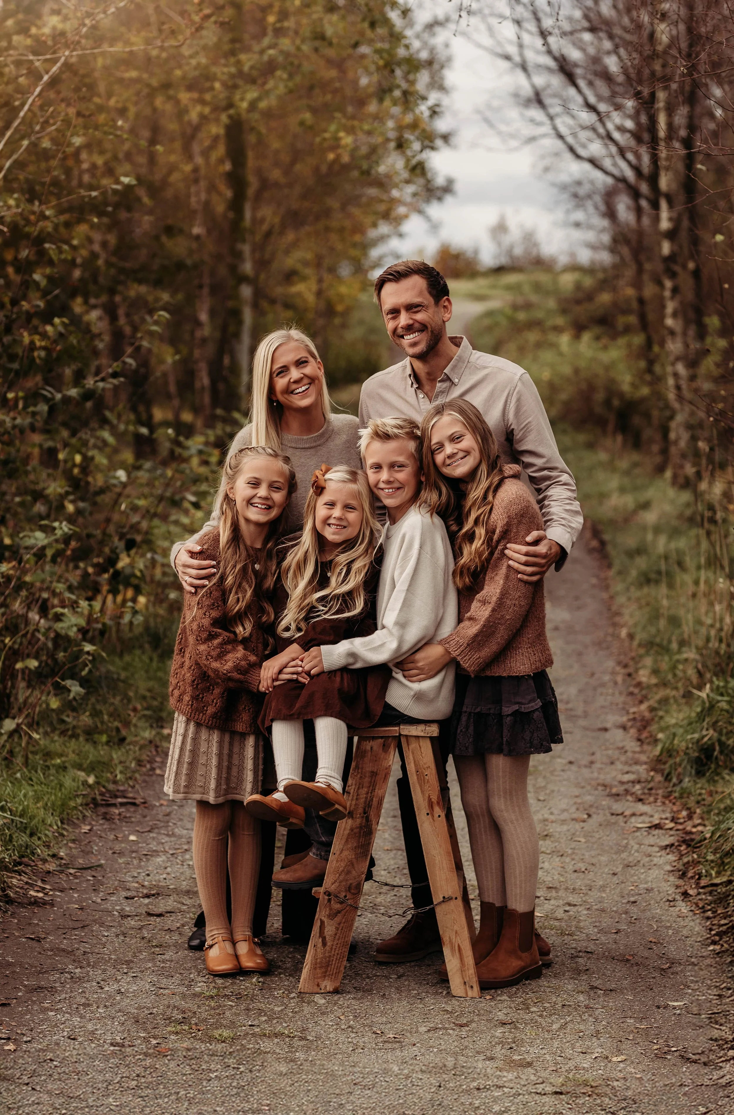 En lykkelig familie på fem, bestående av to voksne og tre jenter, poserer sammen på en tresti i en skog om høsten.