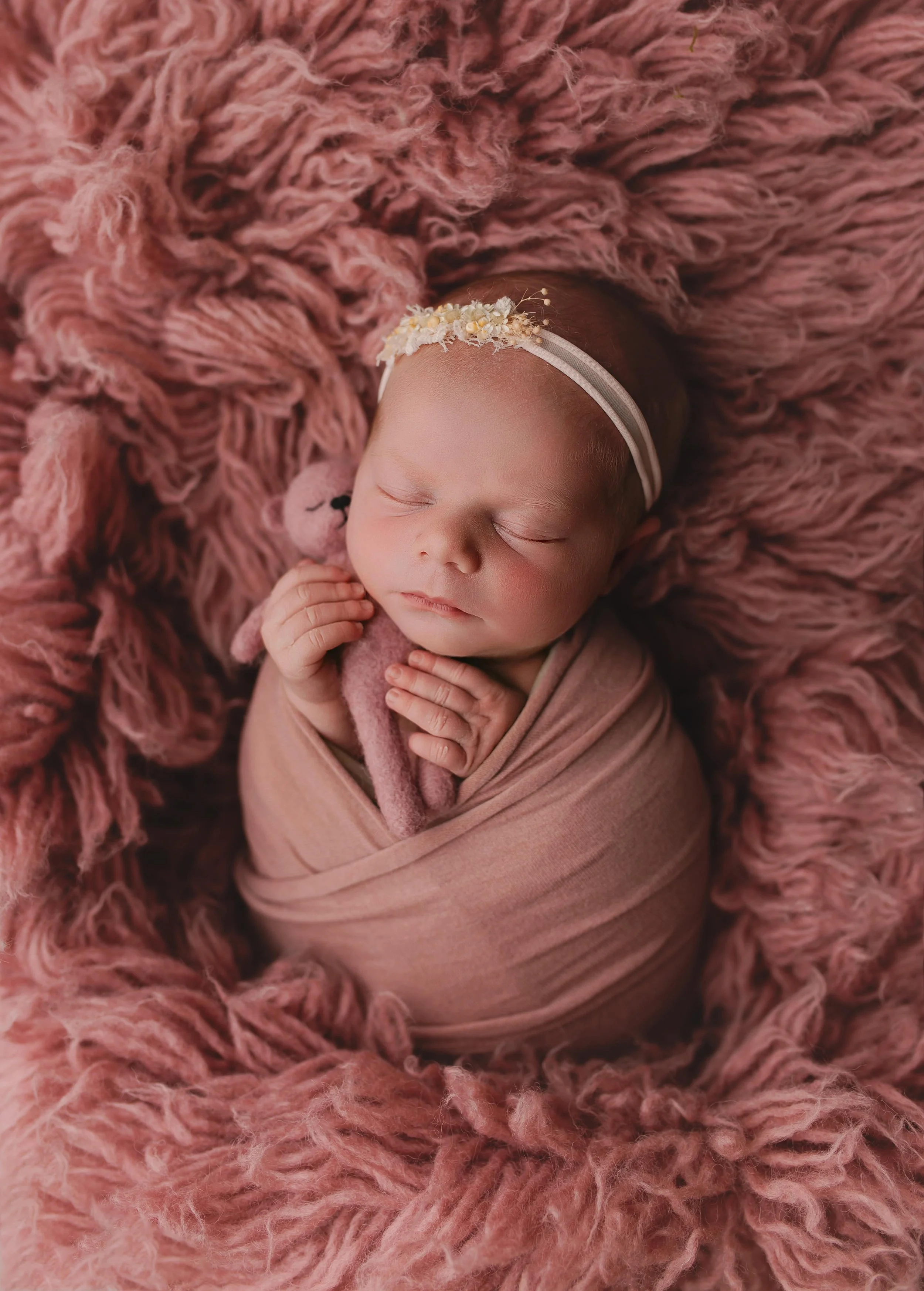 En sovende baby er pakket inn i et lyserødt klær og ligger på en rosa fluffy seng, holder en liten strikket bjørn og har en blomsterhodebånd.