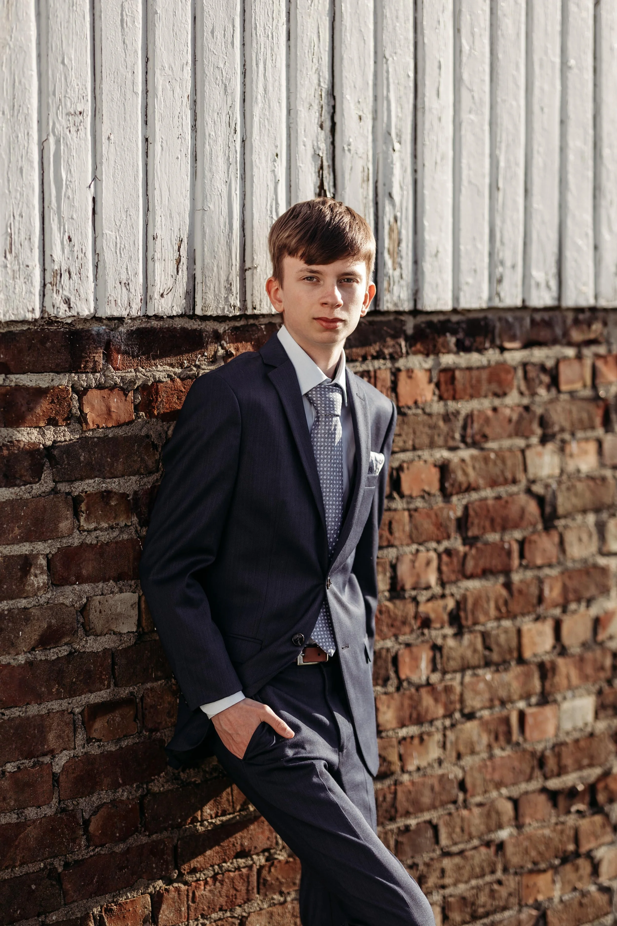 Ung mann i dress står mot en murvegg, med håret til høyre og har en selvsikker uttrykk.