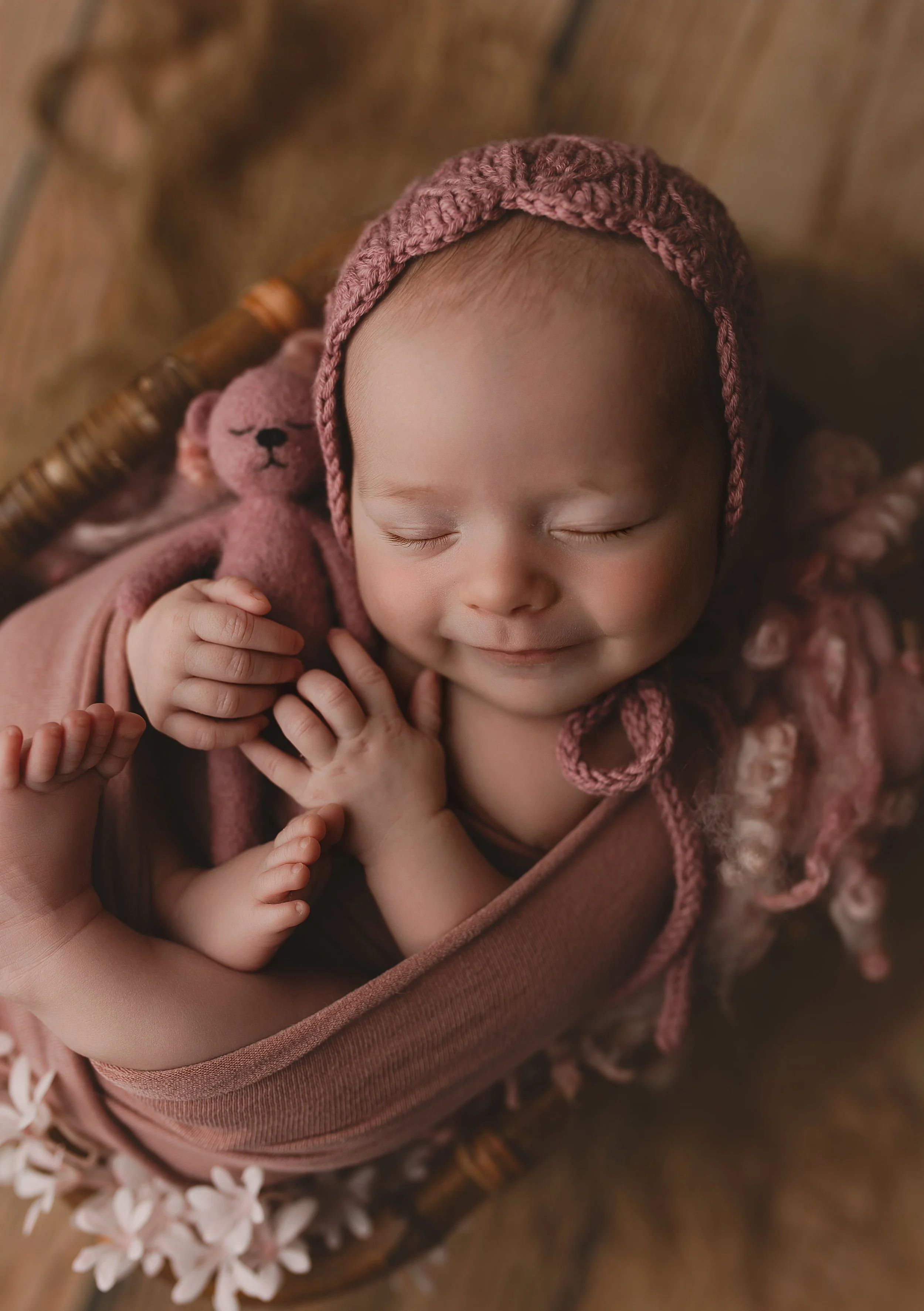 Søvnende baby med rosa hetta og klede, omgitt av en liten rosa bamse i en kurv.