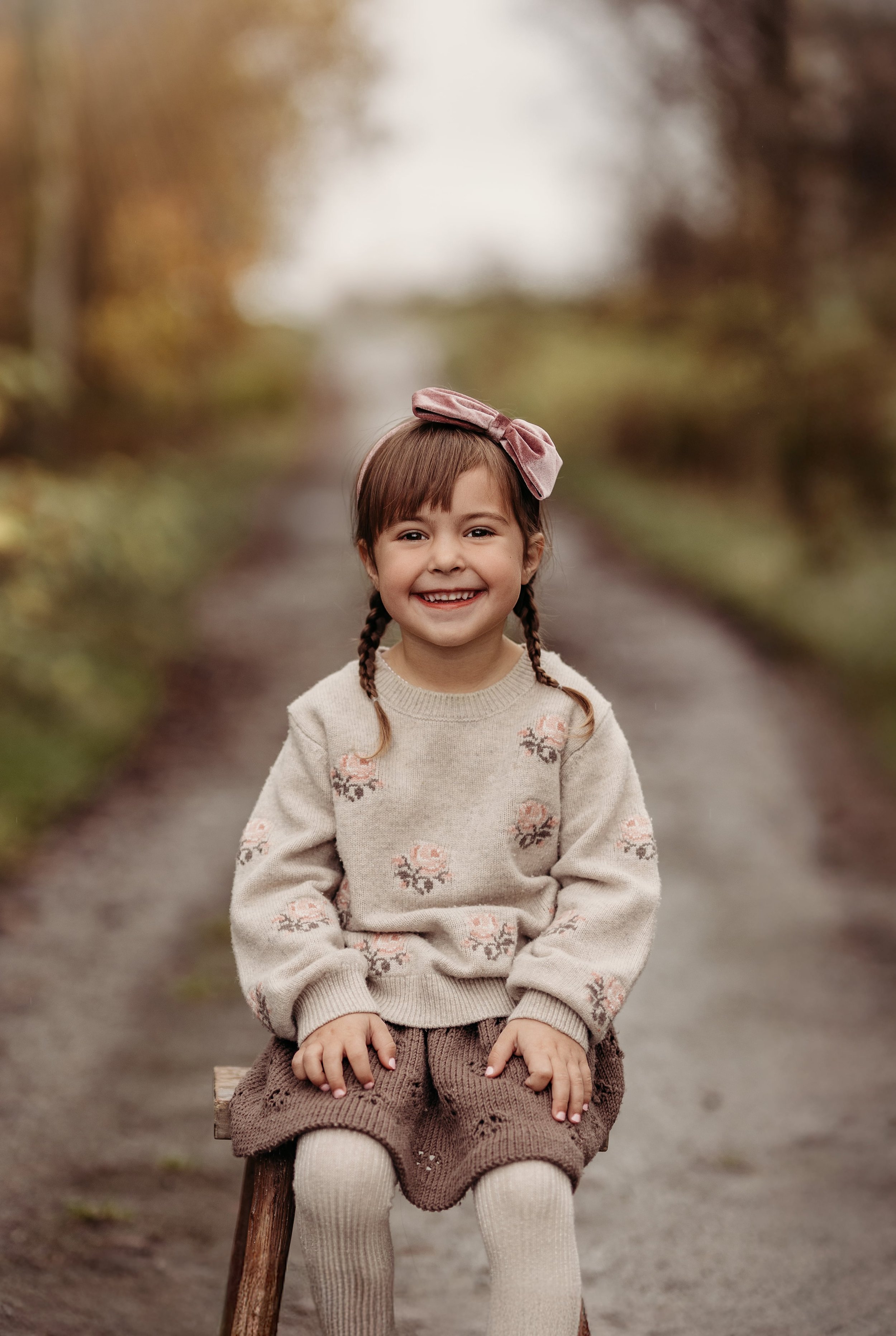 En smilende ung jente med brunt hår og pigtails, iført beige genser med rosaplater og brune bukser, sitter på en liten stol på en sti i en skog om høsten.