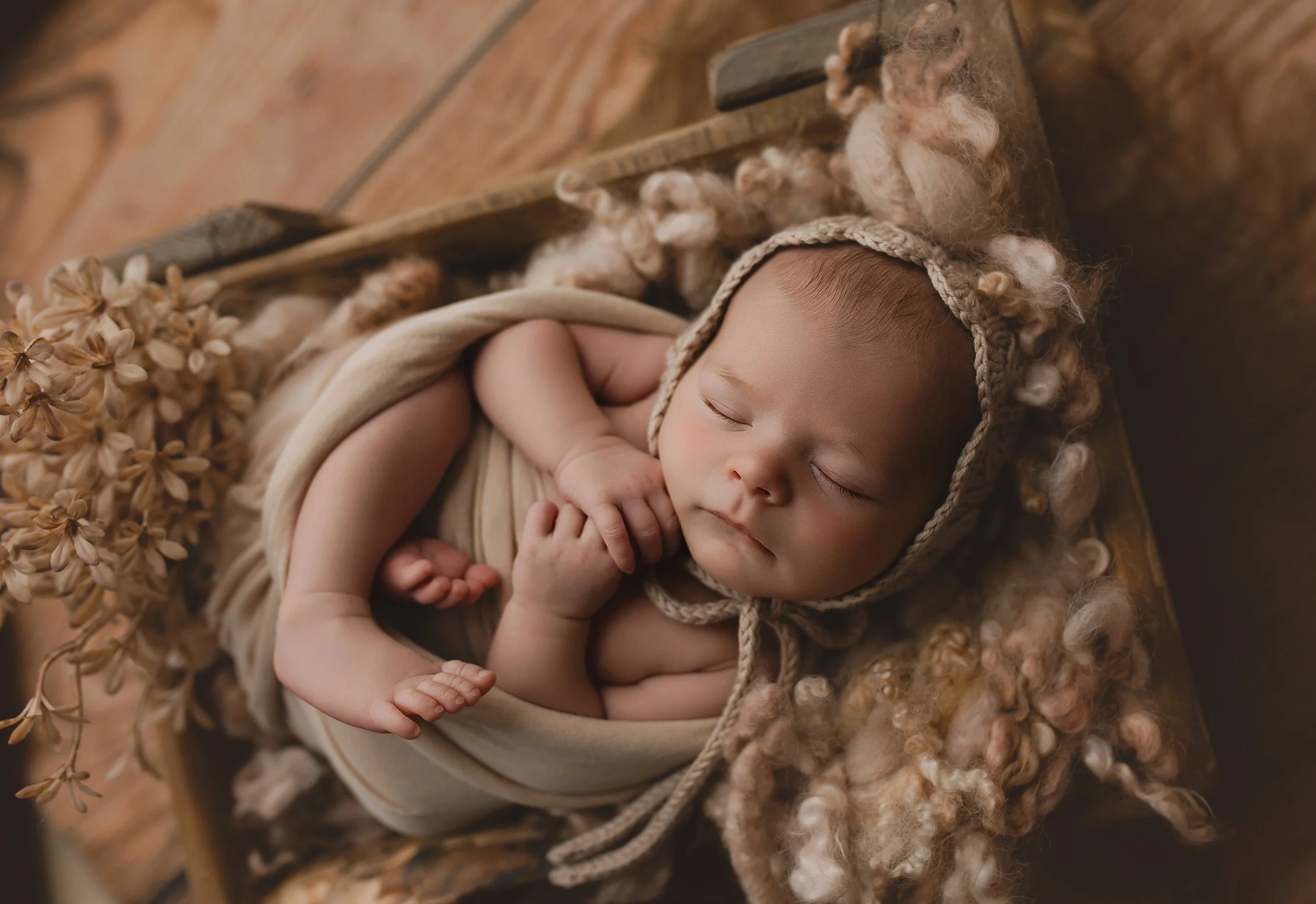 Nydelig baby sovende med krøllete hår, pakka inn i beige teppe, liggende på en trekrakk med tørkede blomster og ullaktig materiale rundt.