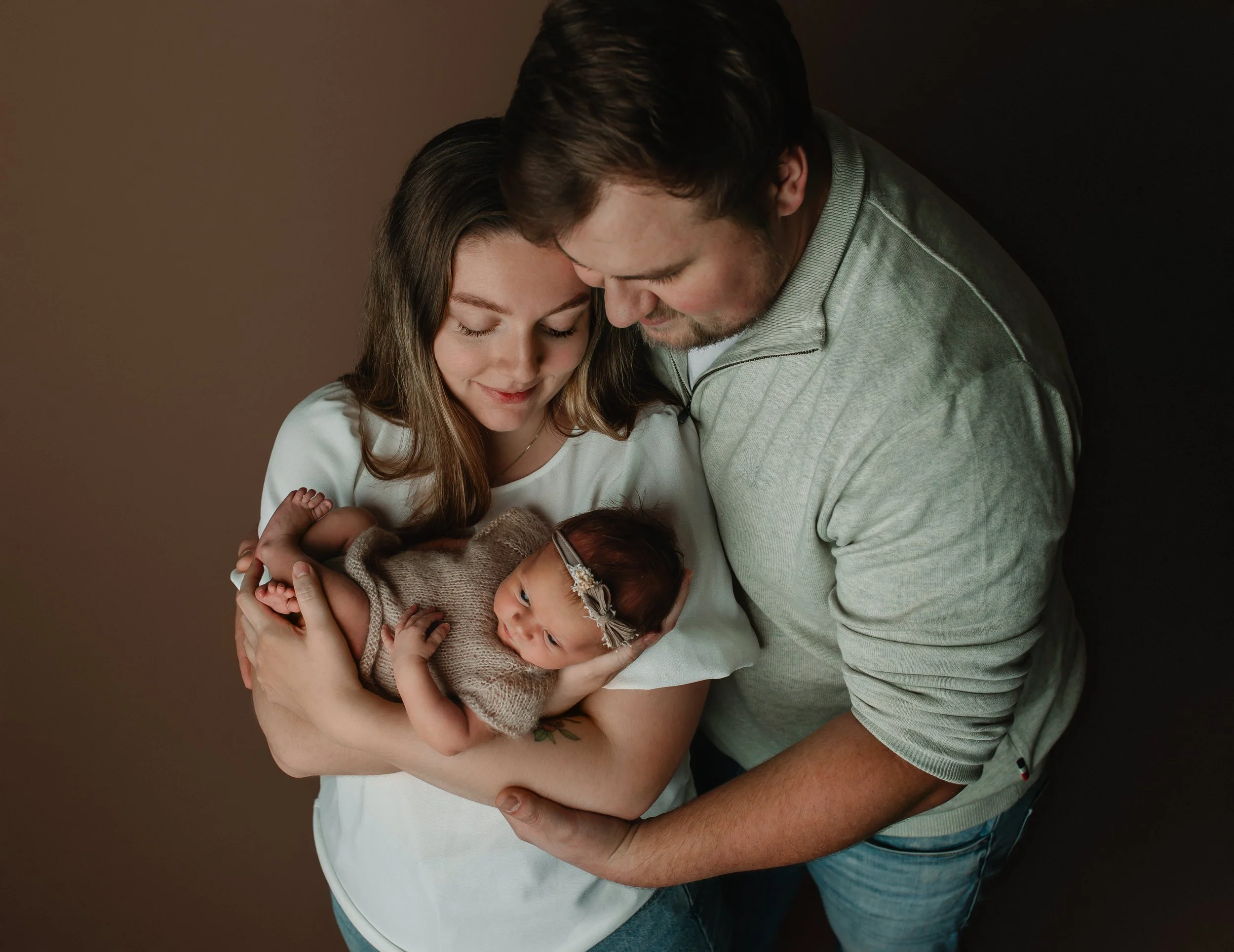 Et ungt par holder et nyfødt barn i armene, de smiler og ser ned på babyen. Bakgrunnen er enkel og mørk.
