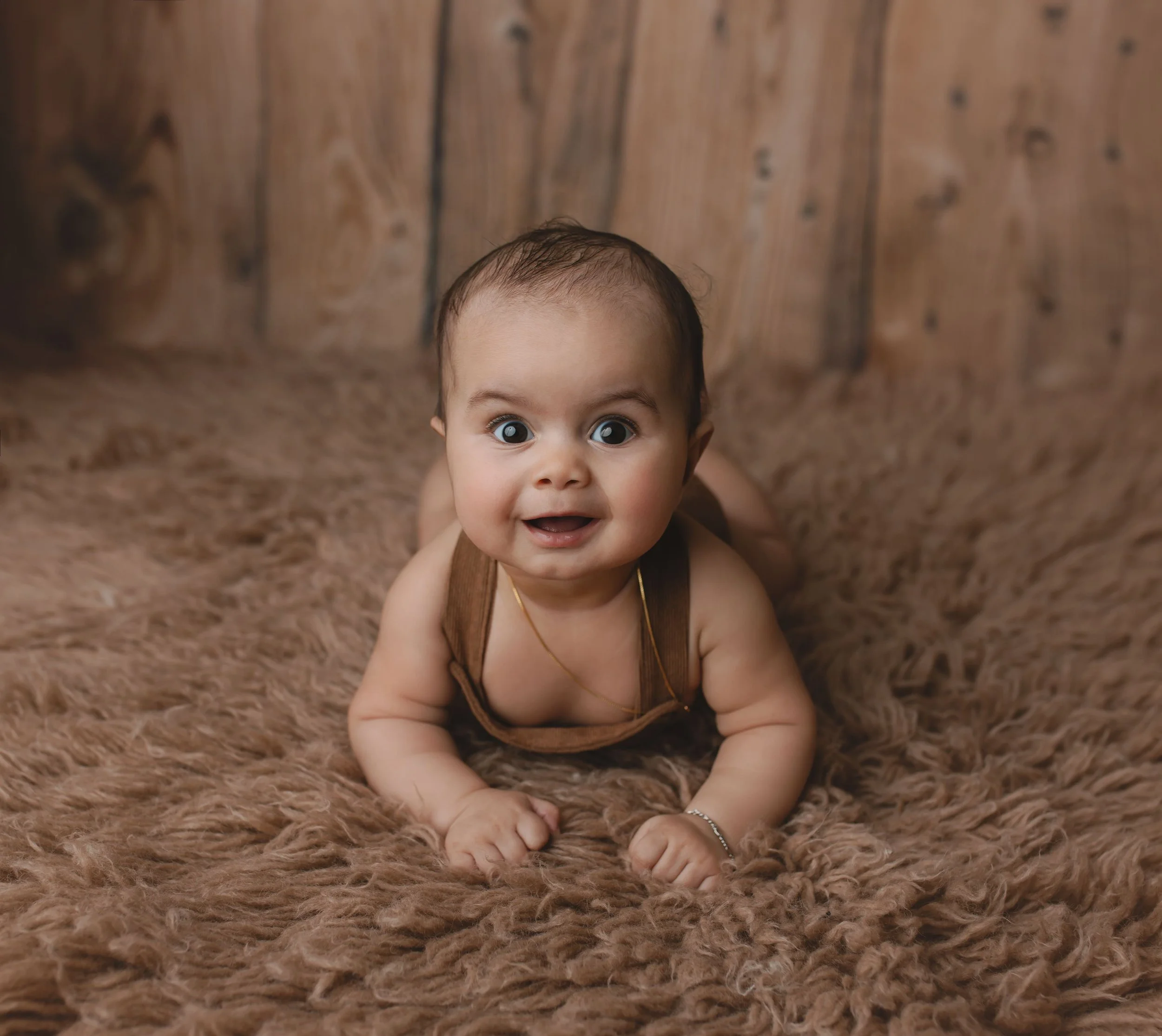 En baby som ligger på magen på en brun, pelslignende matte foran en trevegg.