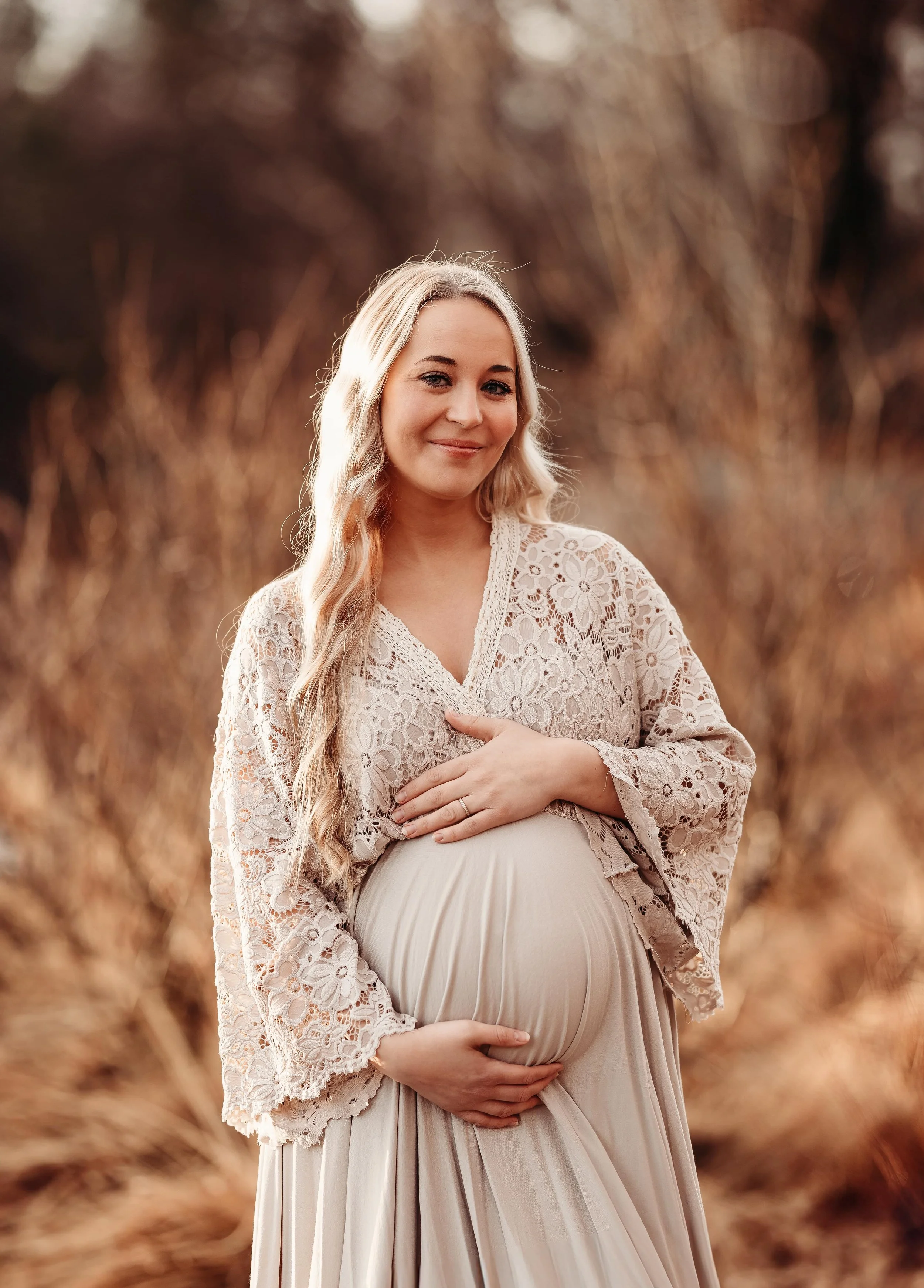 En gravid kvinne i en beige kjole og en blondest bluse, som holder magen sin med begge hender, står i en skog med høstfarger.