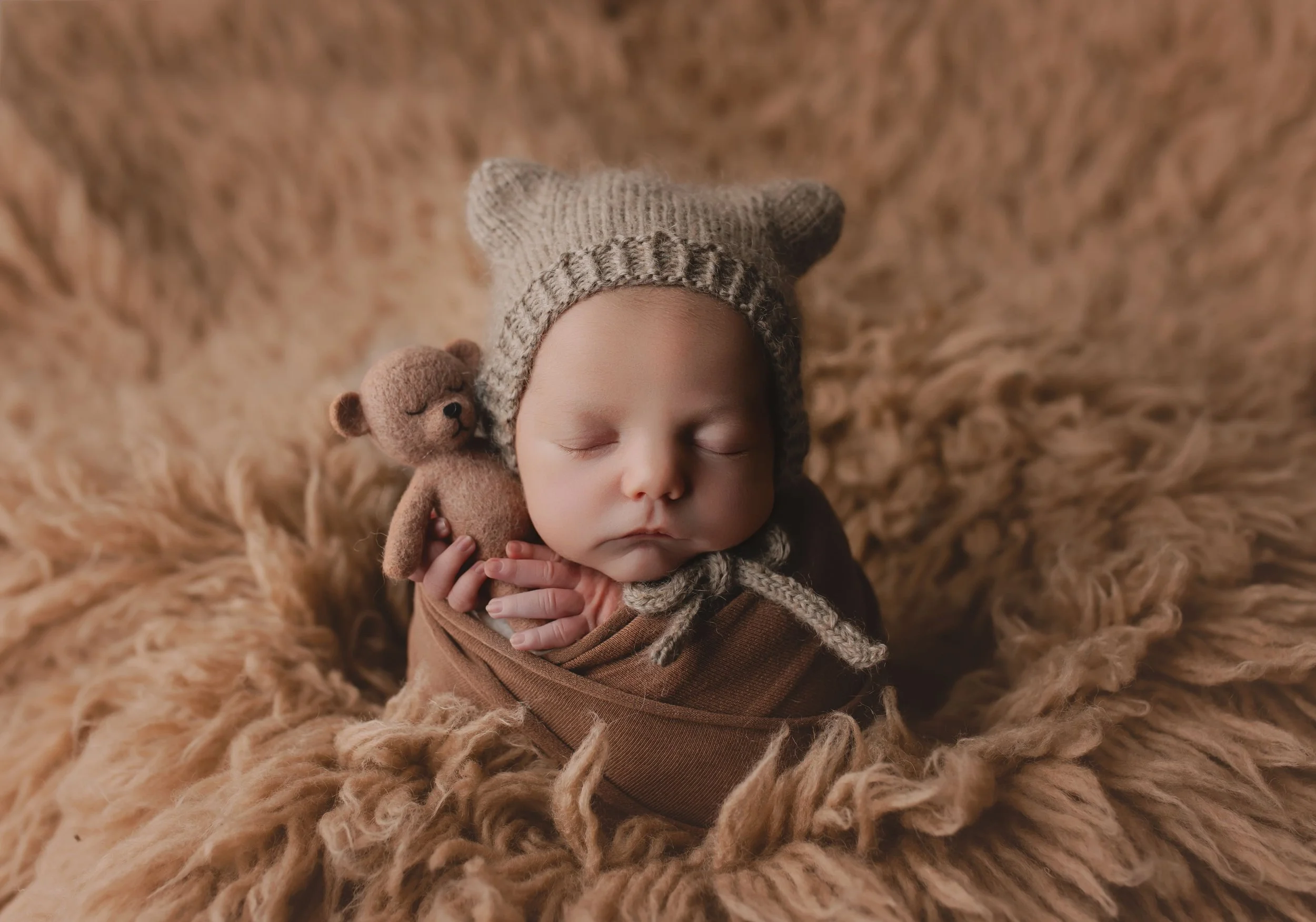 En sovende baby med knapp nesebor, lukkede øyne og lyshår, iført en beige og brun lue med knytinger, som holder en liten brun bamse. Babyen ligger på en myk, beige pelsdekken.