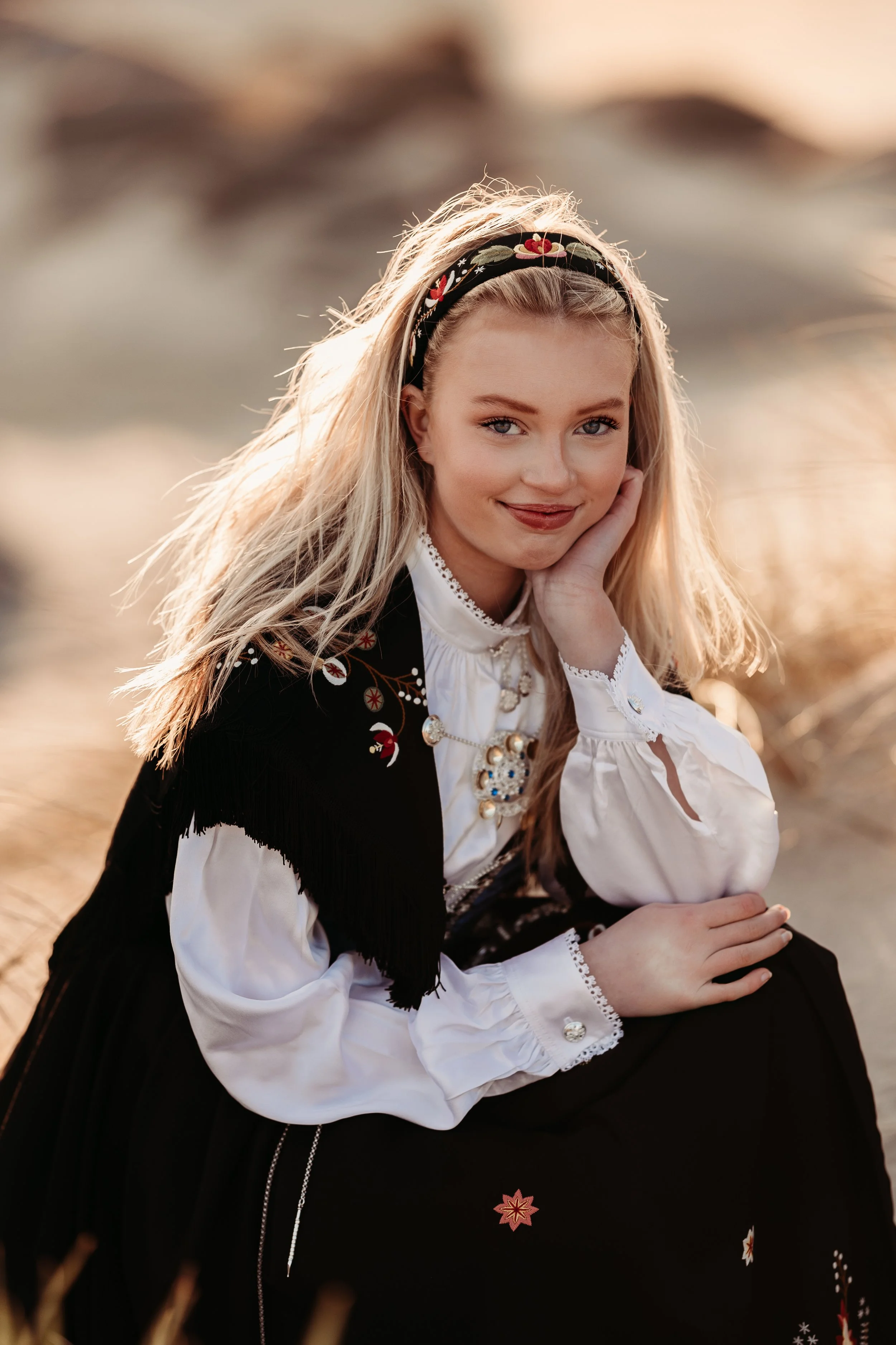 Ung kvinne med langt blond hår iført tradisjonell norsk bunad med broderier, smykker og hårbånd, sitter utendørs i et landskap med solnedgangsskyer.