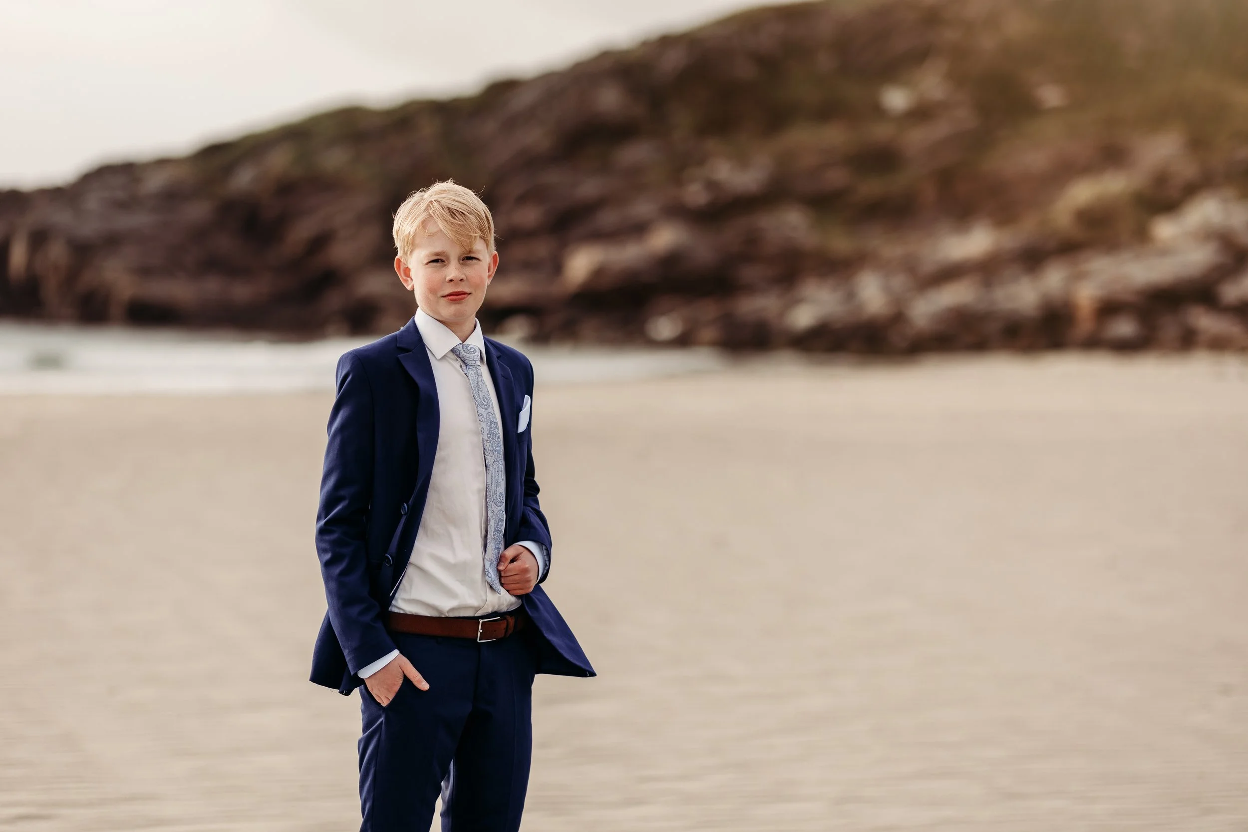 En ung gutt i dress står på en strand med en klippe i bakgrunnen.