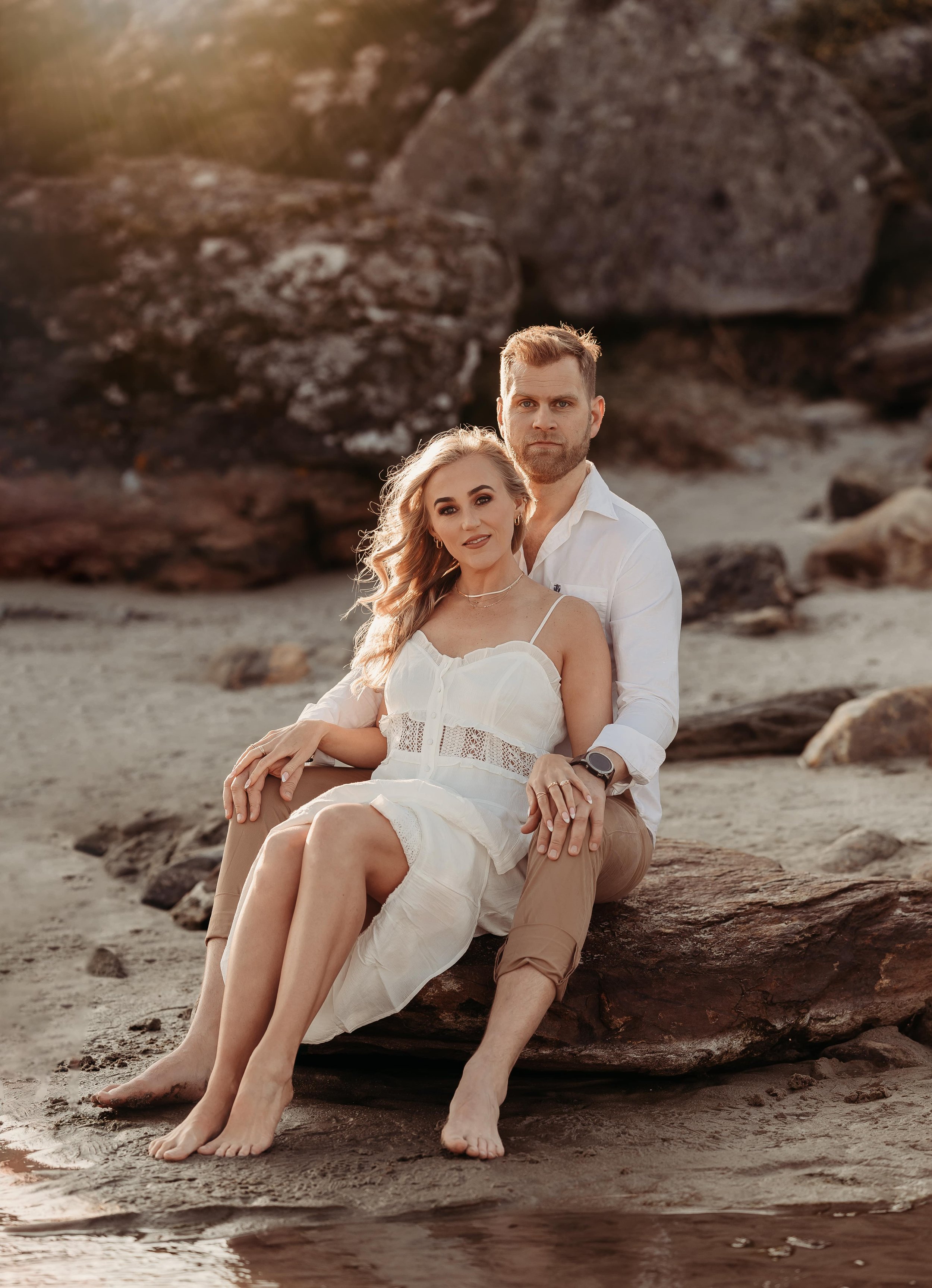 Et par sitter på en stein på stranden, solnedgang bak store steiner i bakgrunnen.