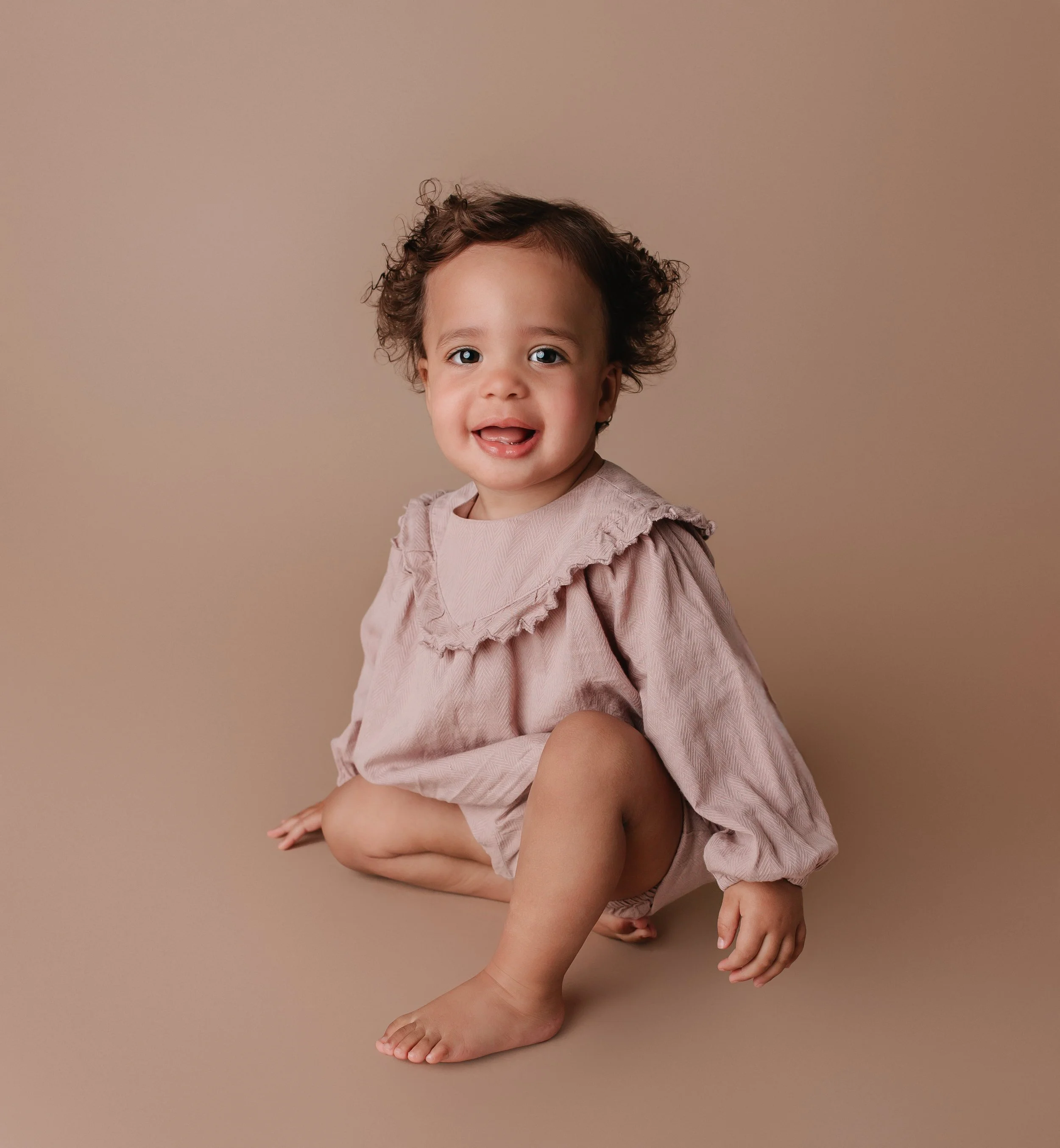Ung jente med krøllet hår, iført rosa kjole med volangkant, som sitter på gulvet mot en beige bakgrunn.