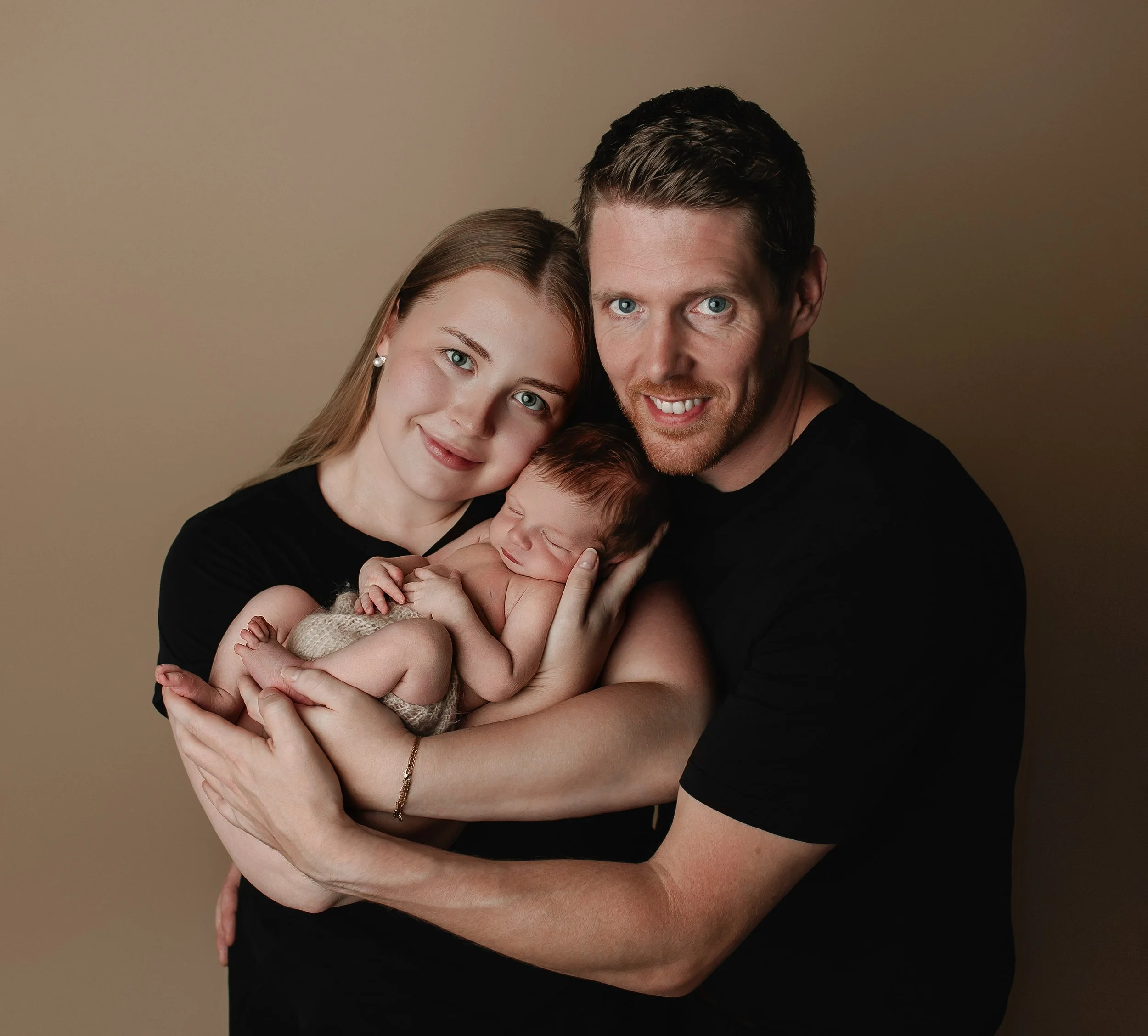 Et lykkelig mor og far holder en nyfødt baby i armene, alle smiler mot kameraet.