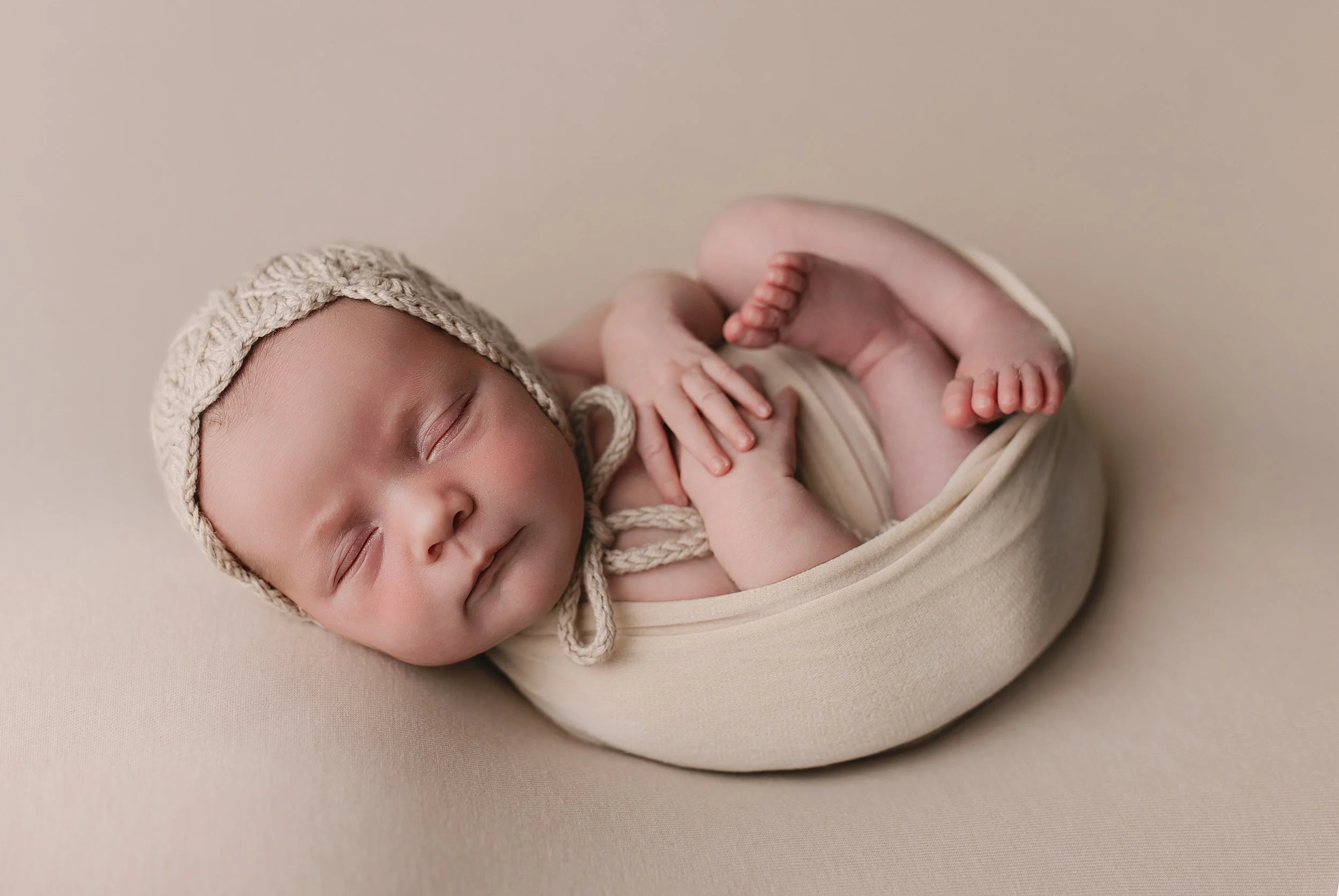 Sovende baby med beigefarget taupe bakgrunn, iført strikket lue og kremhvit drakt, liggende i svøpe.