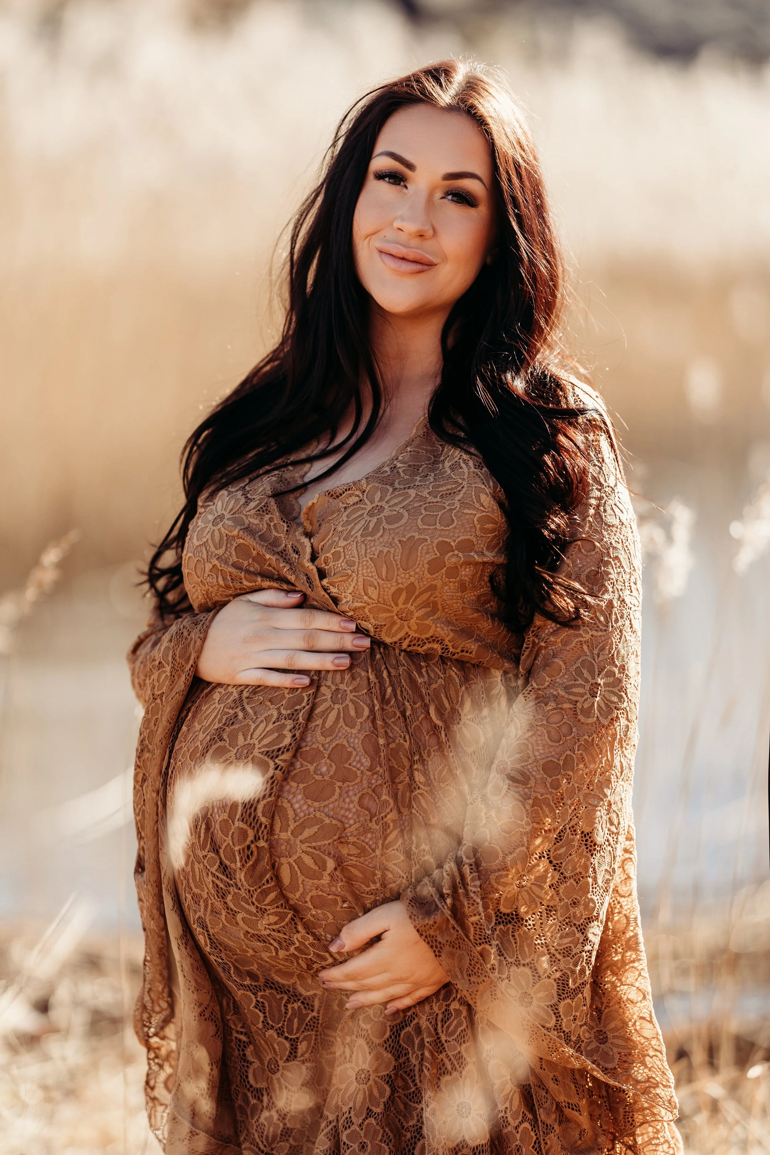 En gravid kvinne som holder magen sin i en ute i solen, iført en brun blonder kjole. Bakgrunn med gress og sollys.