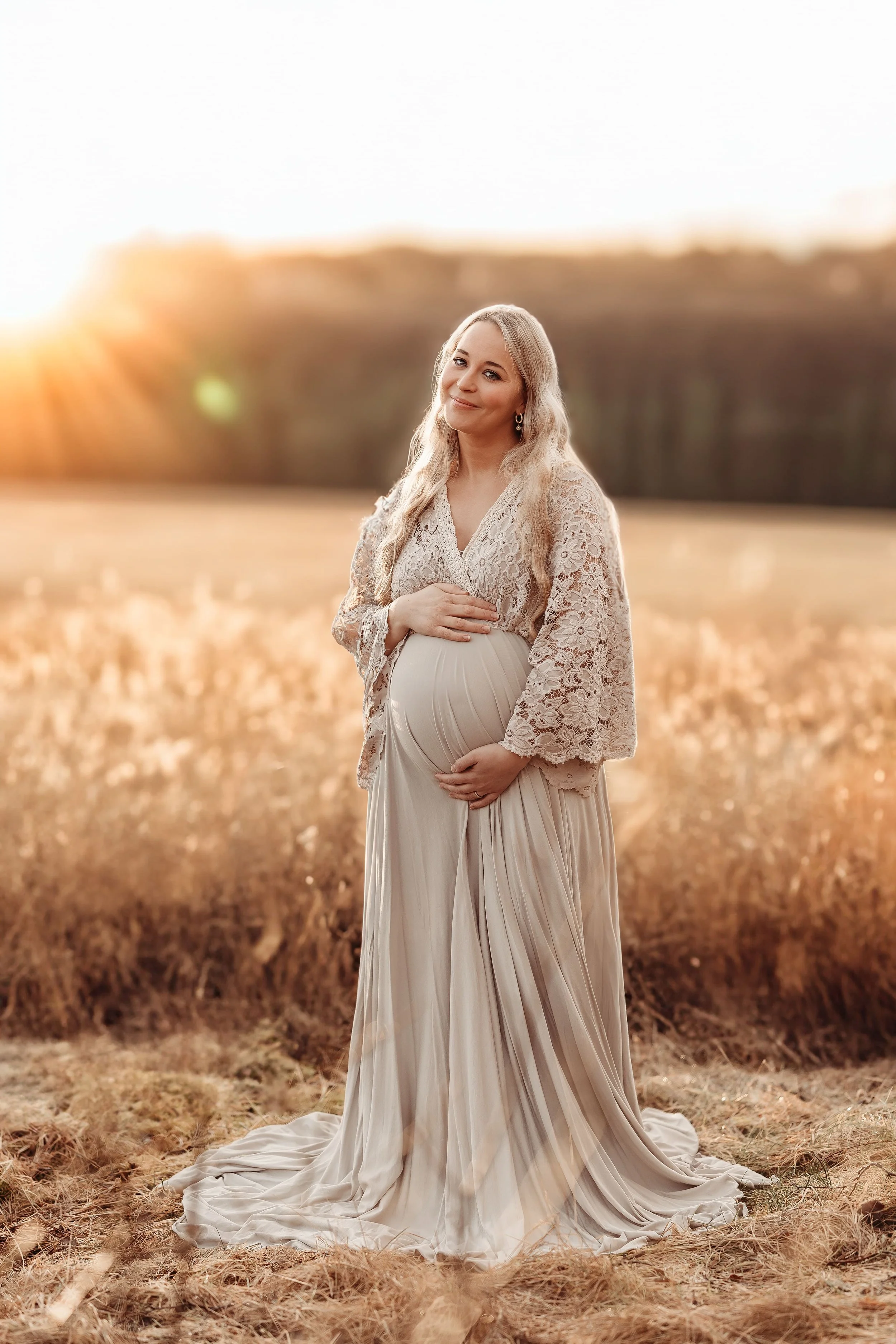 En gravid kvinne kledd i et lyst, flytende kjole med lace-overslag, står i en åpen eng med høstfarger, solnedgang i bakgrunnen.