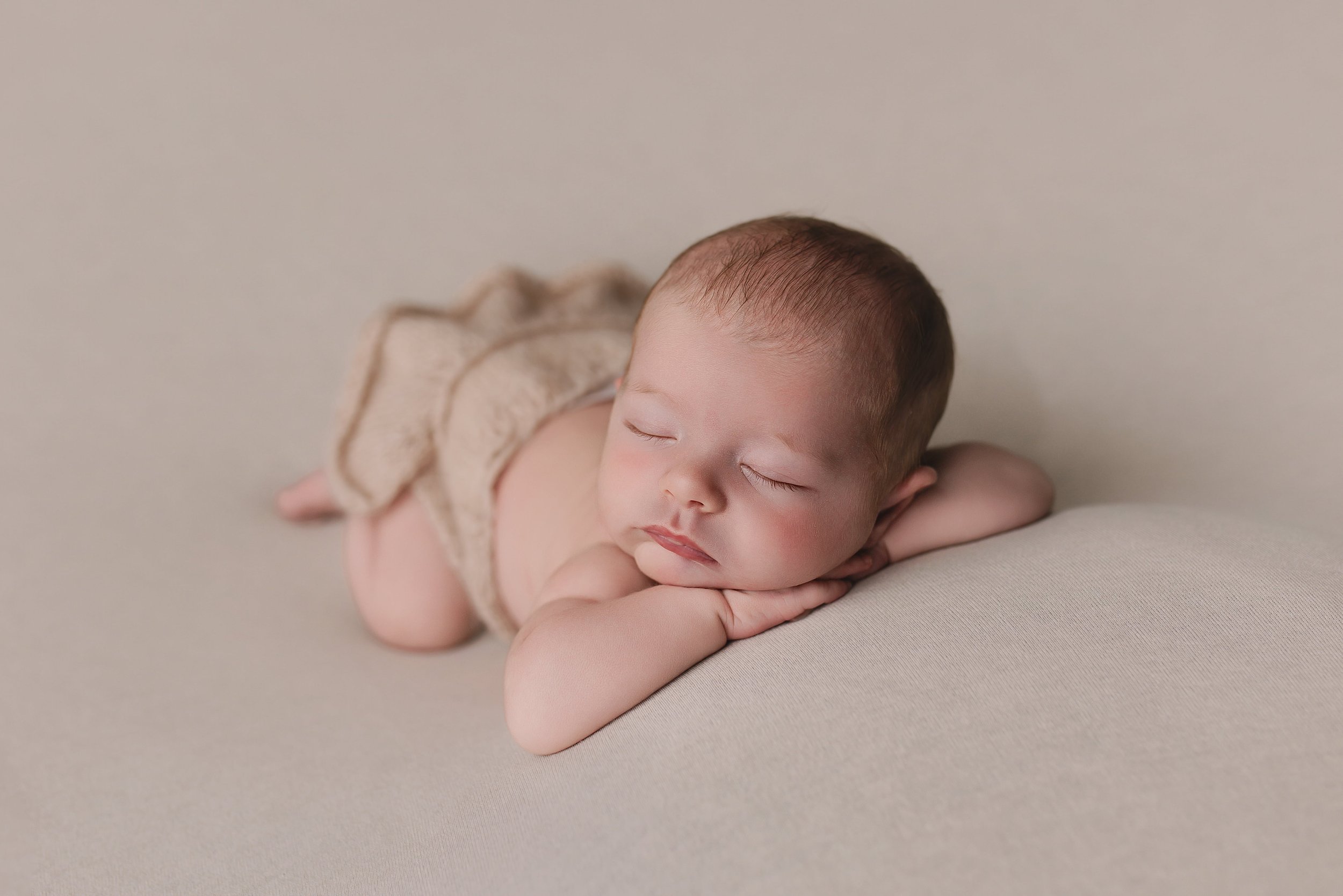 En baby ligger på magen på en myk, lysfarget overflate med øynene lukket og en rolig ansiktsuttrykk.