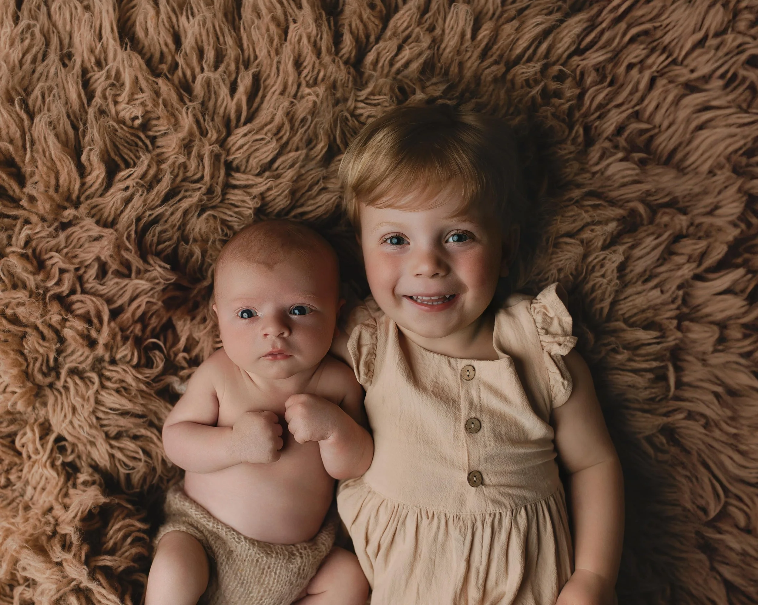 En liten baby og en ung jente ligger på en brun, fluffy plaid. Babyen er uten klær og holder hendene foran brystet, mens den unge jenta smiler og har på seg en beige kjole med puffede ermer.