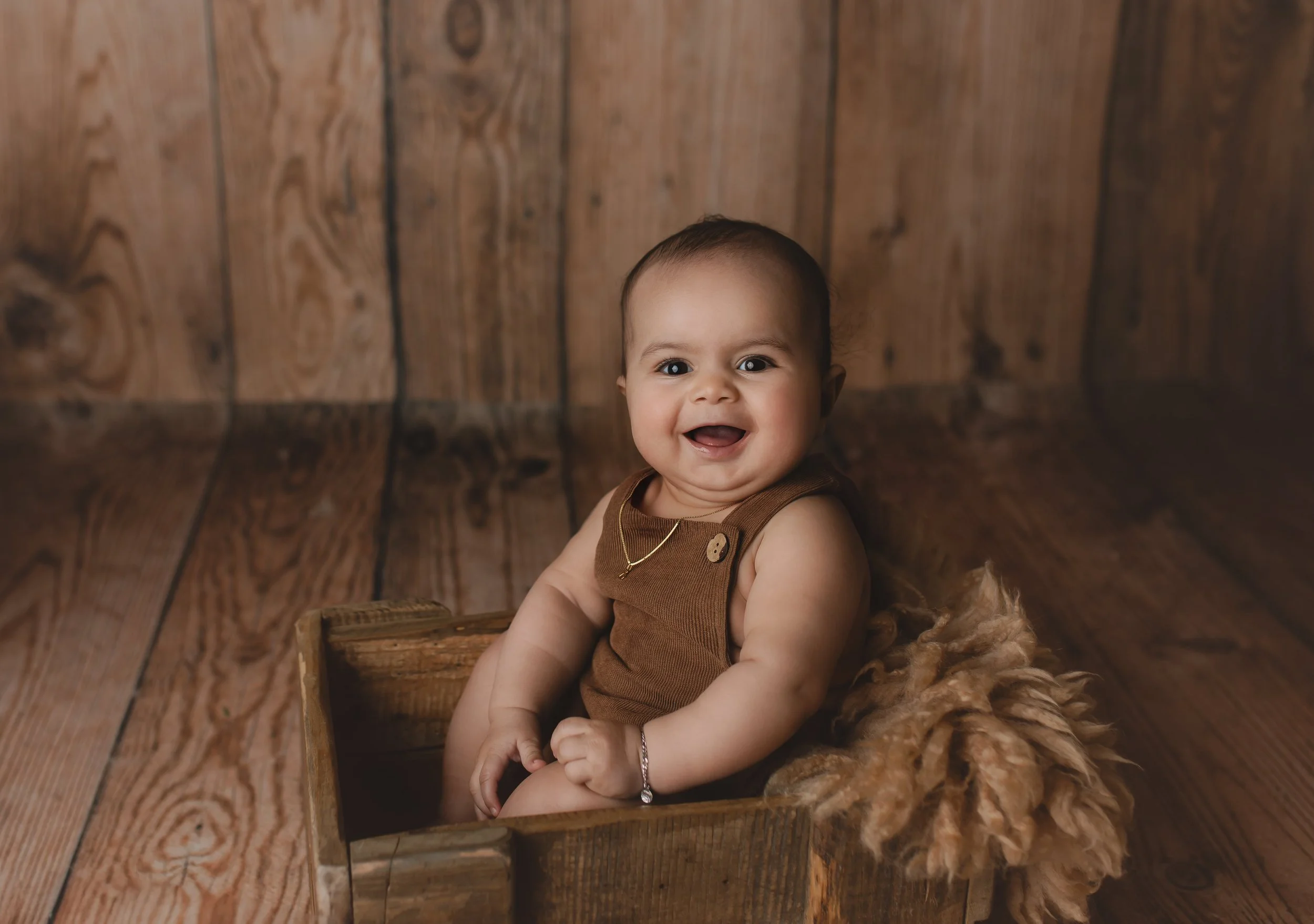 En smilende baby i en brun overdel som sitter i en treboks, omgitt av en myk, brunsfarget teppe, mot en bakgrunn av trebunn og tremplanker.