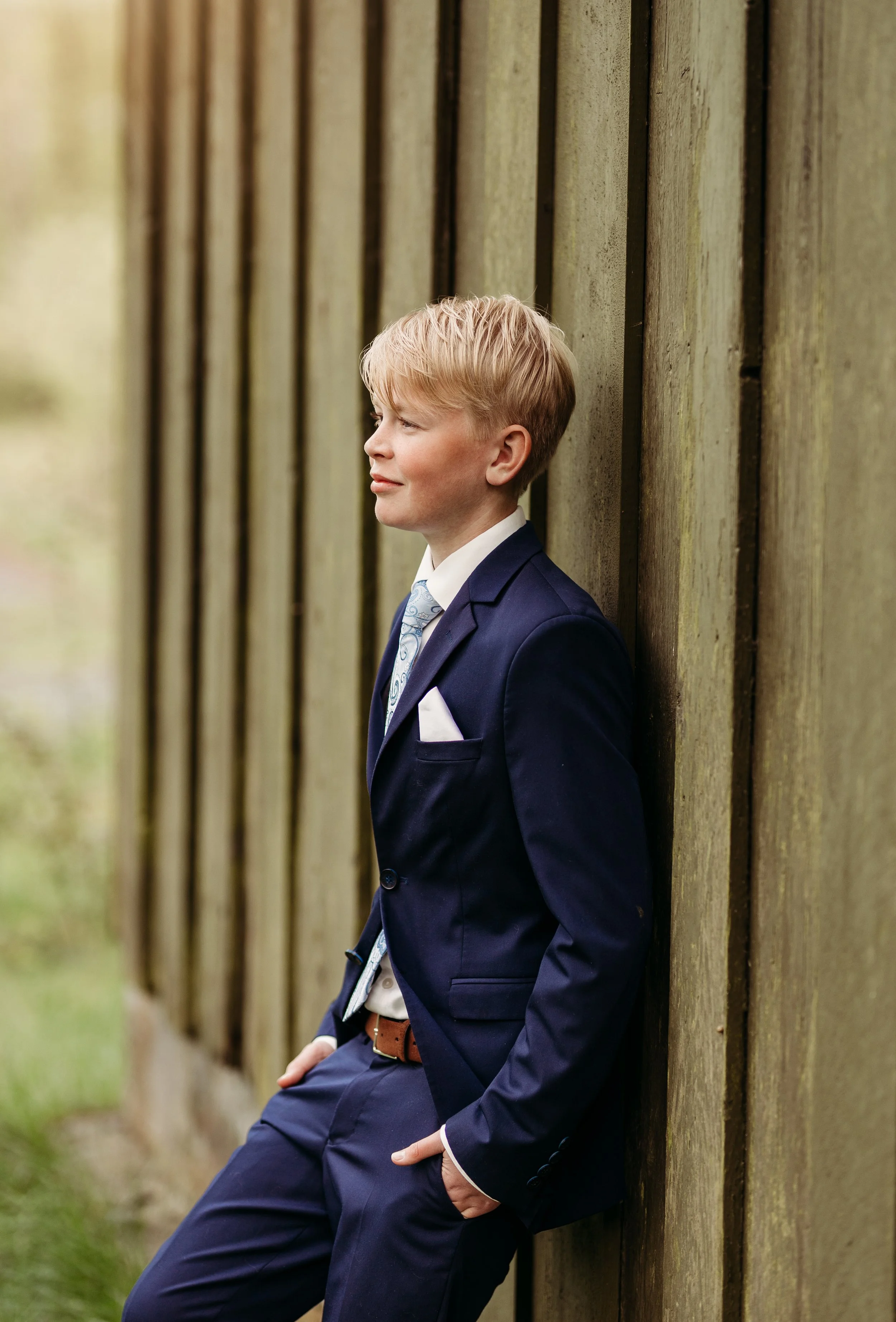 En ung gutt i dress står ved en trefence og ser ut mot venstre.