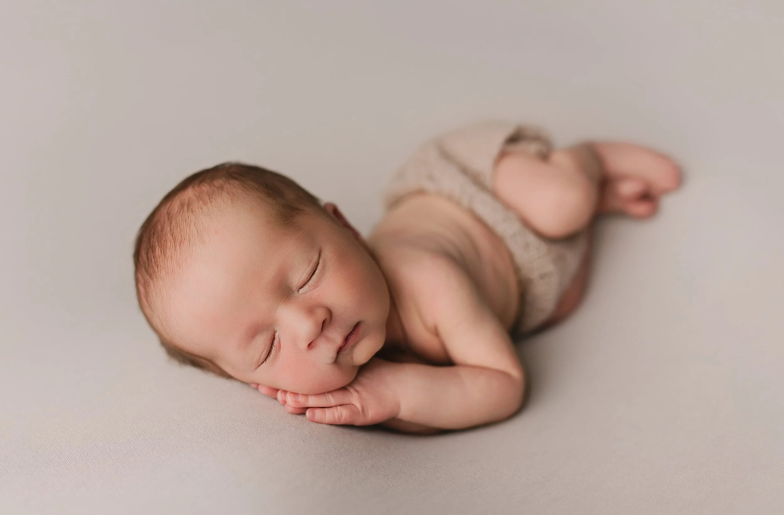 Nybakt baby ligger på siden med hodet hvile på hånden, iført bleie og sover på en lys bakgrunn.