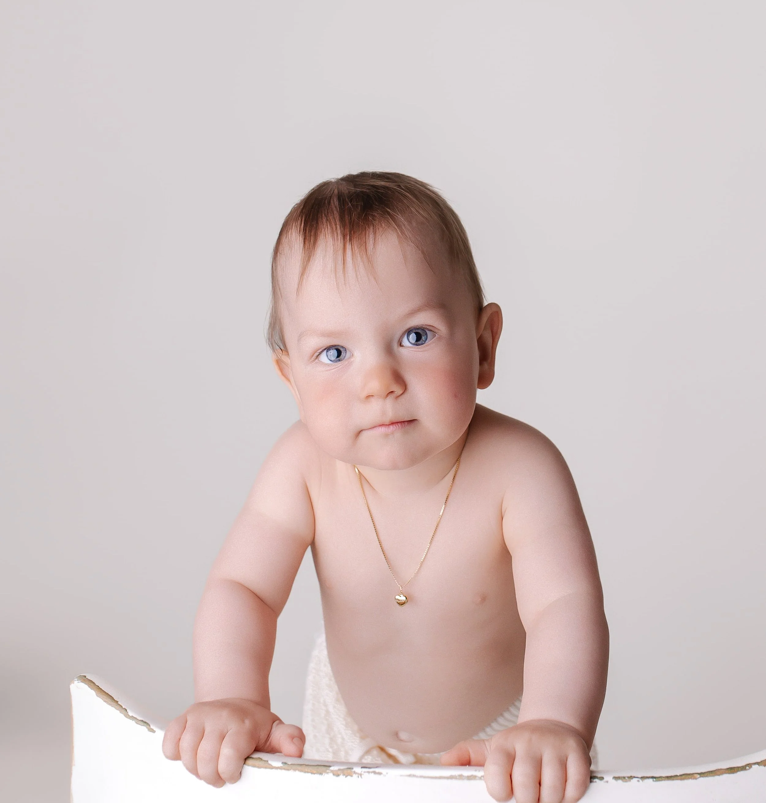 Barnerunge med blå øyne og kort brunt hår som står opp, uten klær bortsett fra en gull halskjede, holder på en hvit overflate mot en lys bakgrunn.
