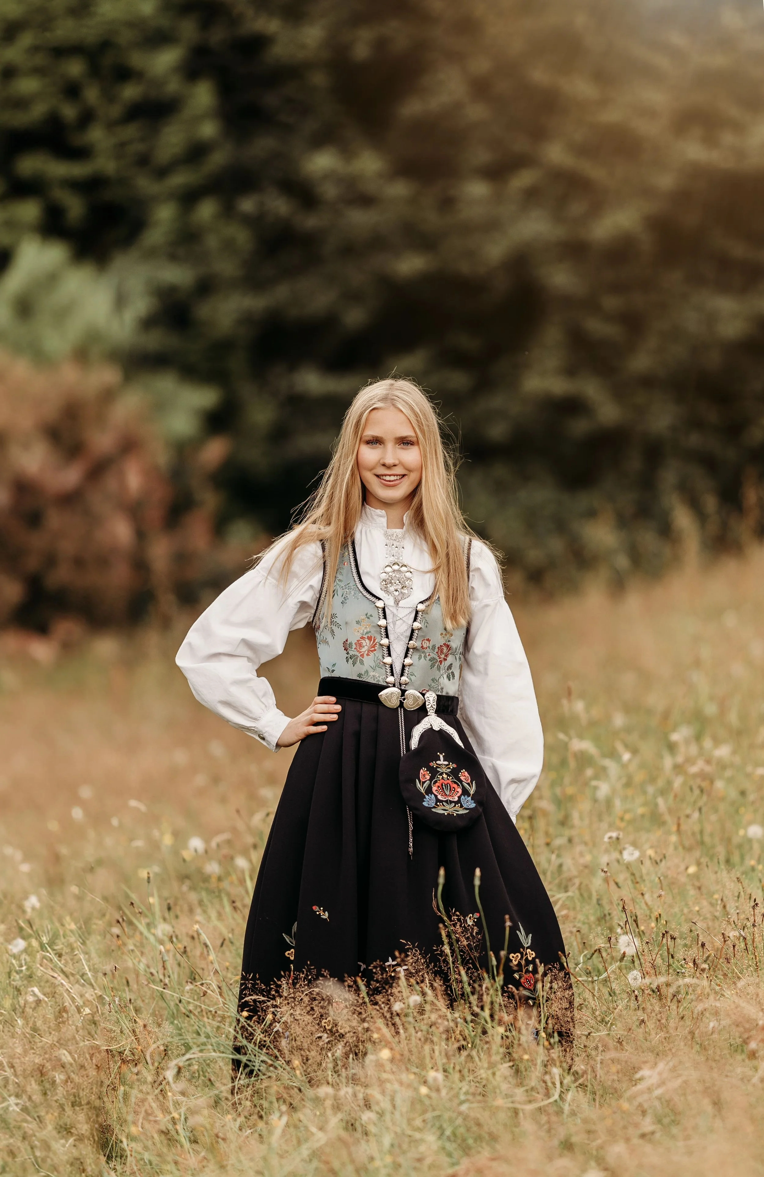 En ung kvinne i tradisjonell norsk bunad står i en eng med gress og blomster, med trær i bakgrunnen.
