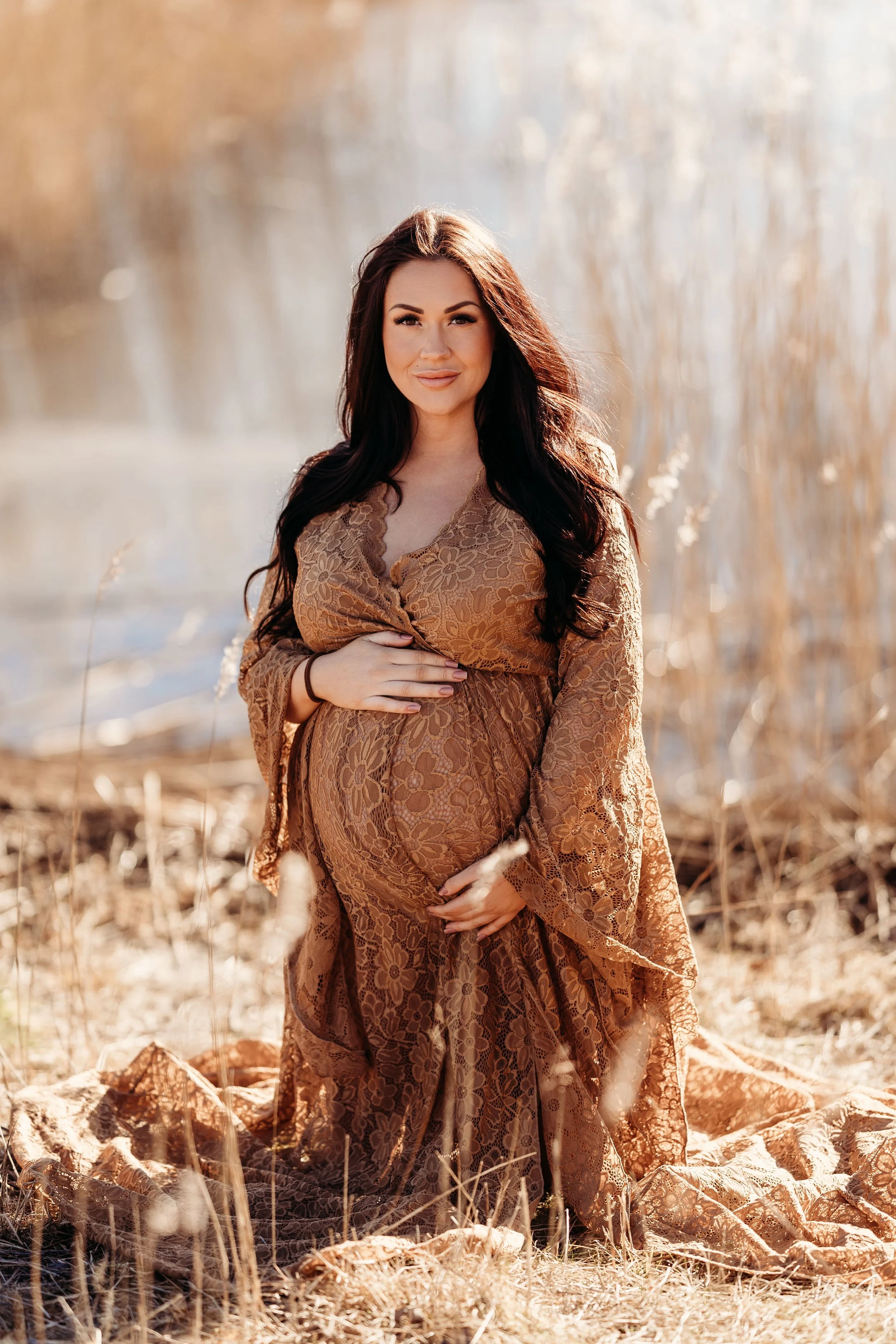 Gravid kvinne i brunt blondekjole står på en tørreng med vann i bakgrunnen.