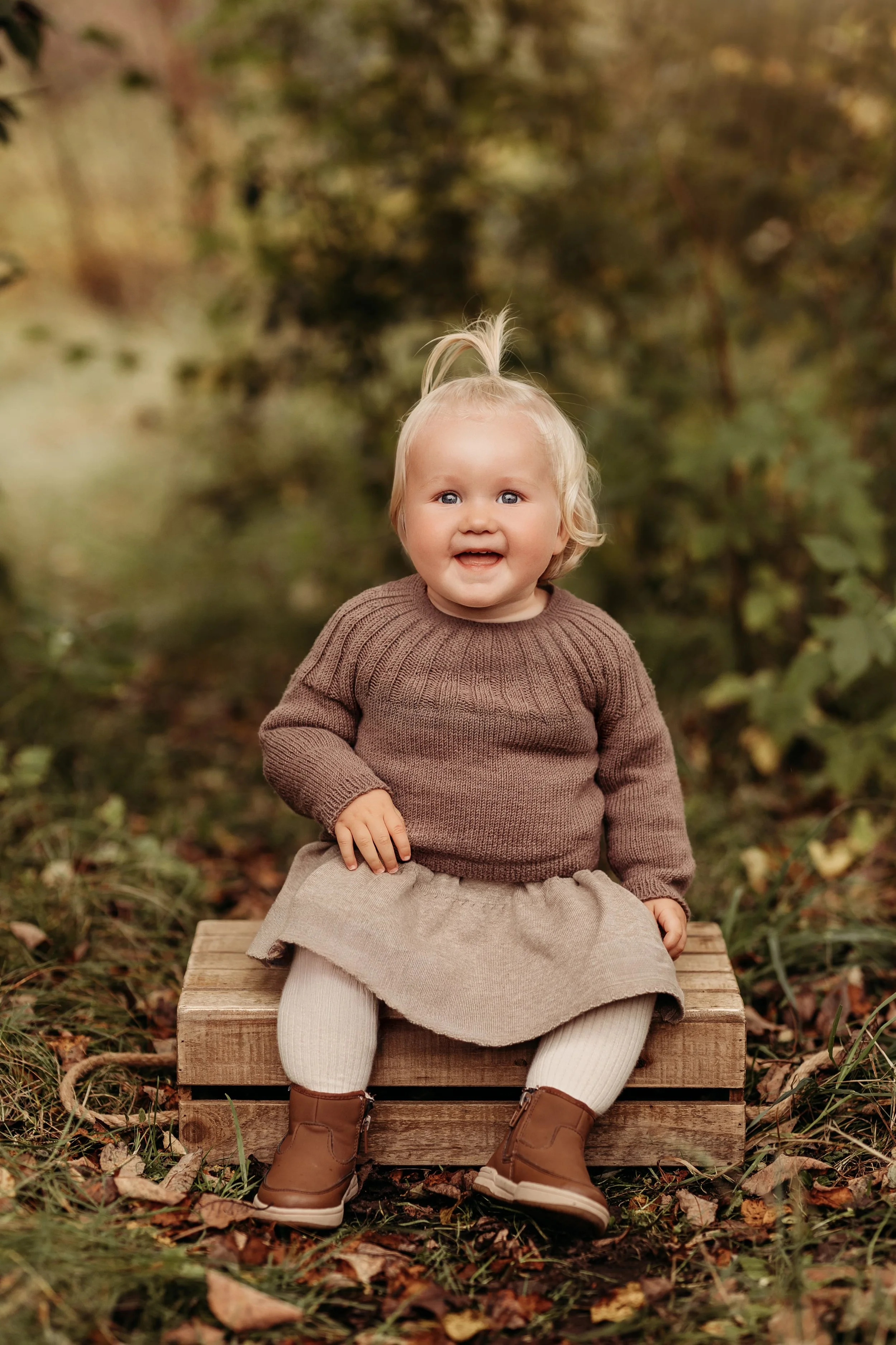 En glad liten jente som sitter på en trekloss i en skog om høsten, iført brun genser, beige skjørt, beige strømper og brune støvler.